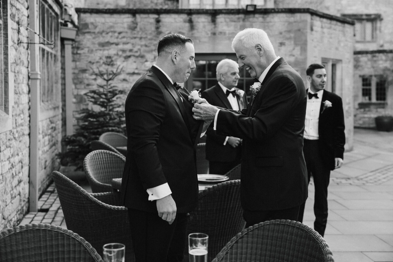 Two groomsmen in suits adjust buttonholes in an outdoor stone courtyard, with other men in formal attire in the background.