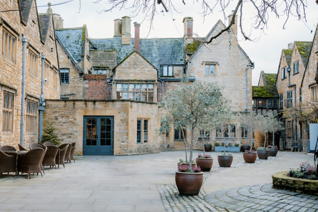 A quaint, stone courtyard of Lygon Arms Hotel with potted plants and wicker chairs arranged around tables, surrounded by historic buildings with pitched roofs and chimneys.