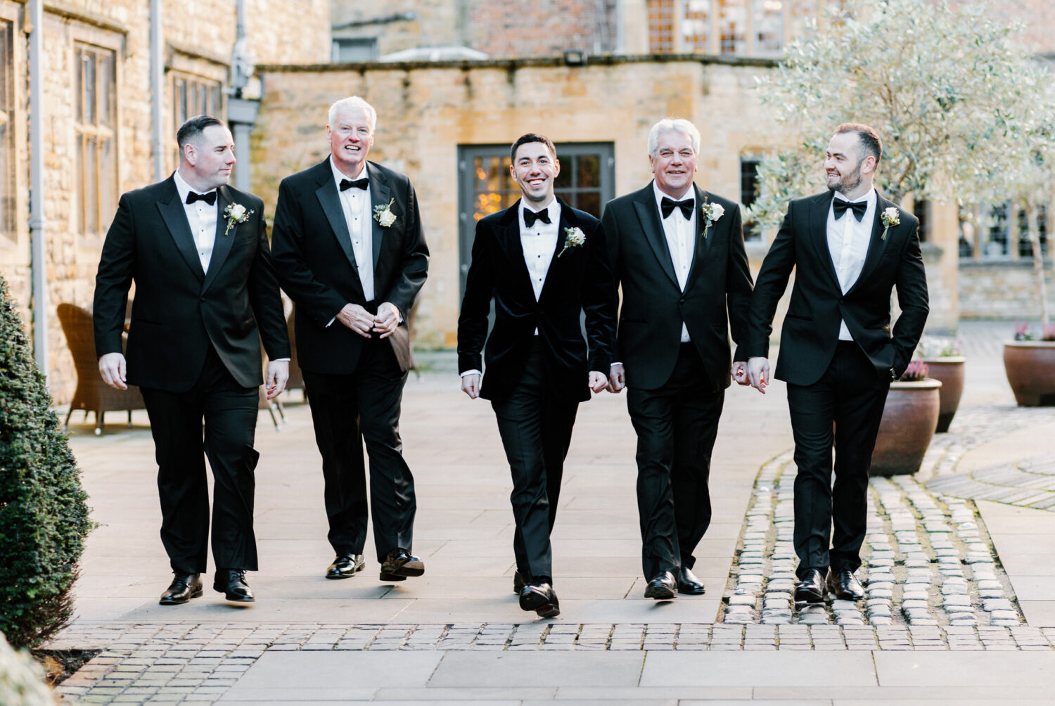 Five groomsmen in tuxedos walk side by side on a cobblestone path outside a building. They are smiling and appear to be in a celebratory mood.