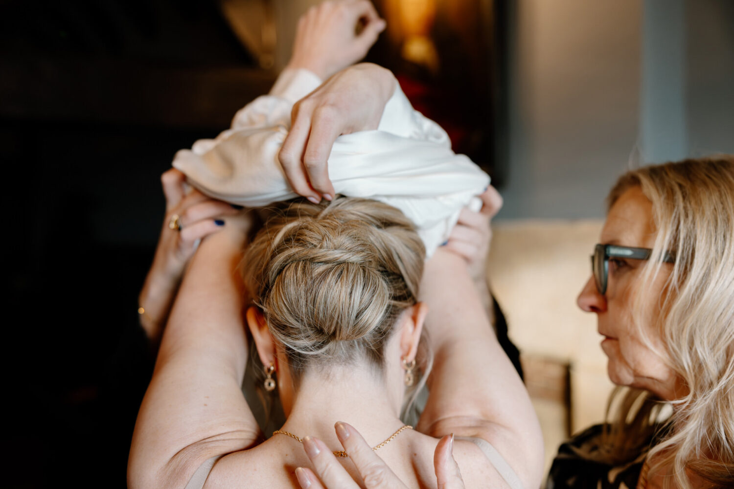 With a blonde updo, the bride helps another person put on a wedding dress, assisted by others.