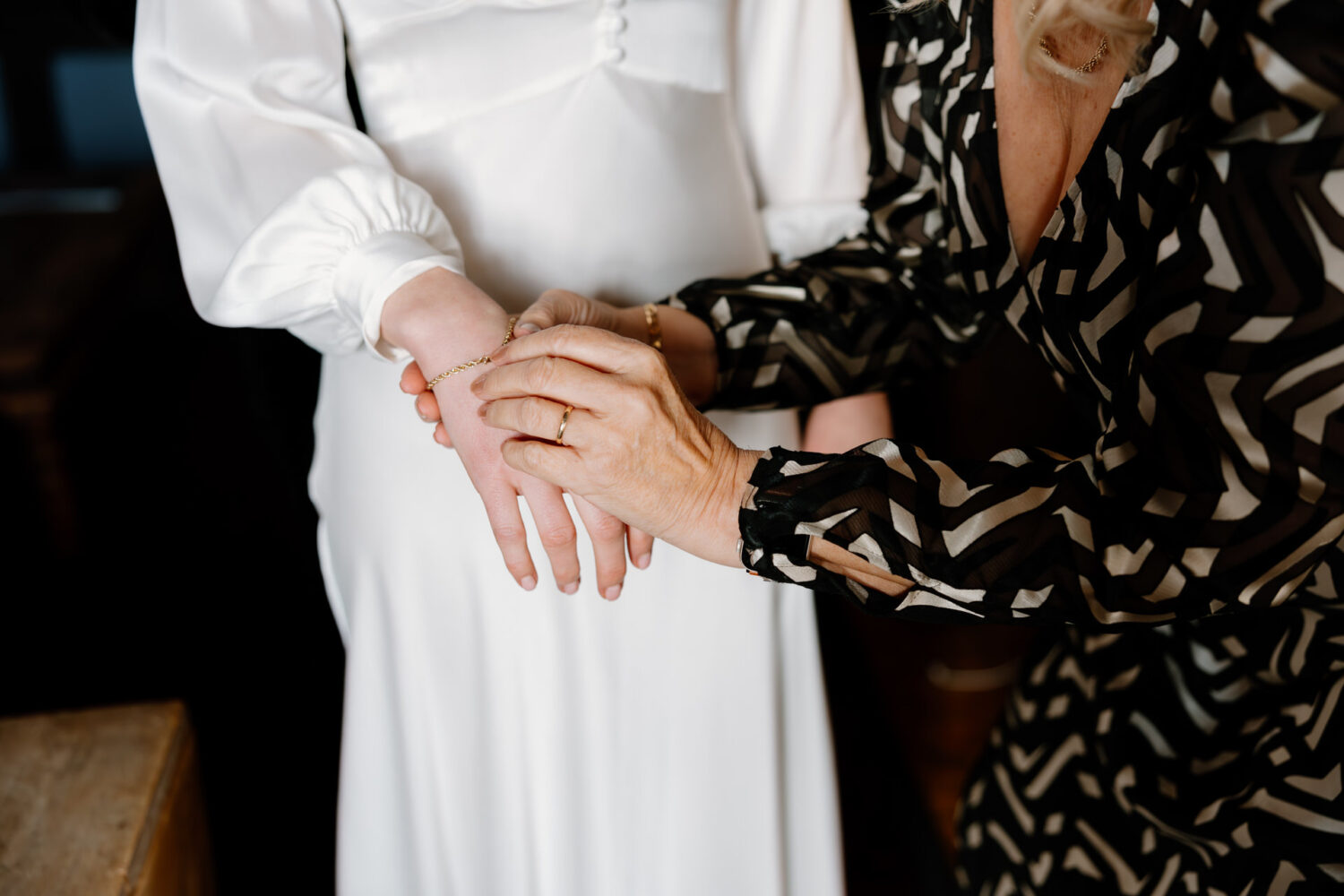 The bride's mother is in a black-patterned dress, fastening a bracelet on another person's wrist and wearing a white dress.