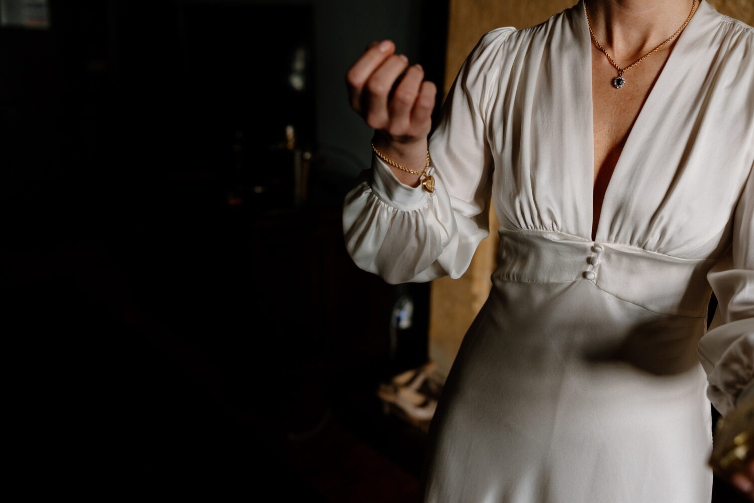 The bride in a white long-sleeve dress, wearing a necklace and bracelet, stands partially visible in dim lighting.