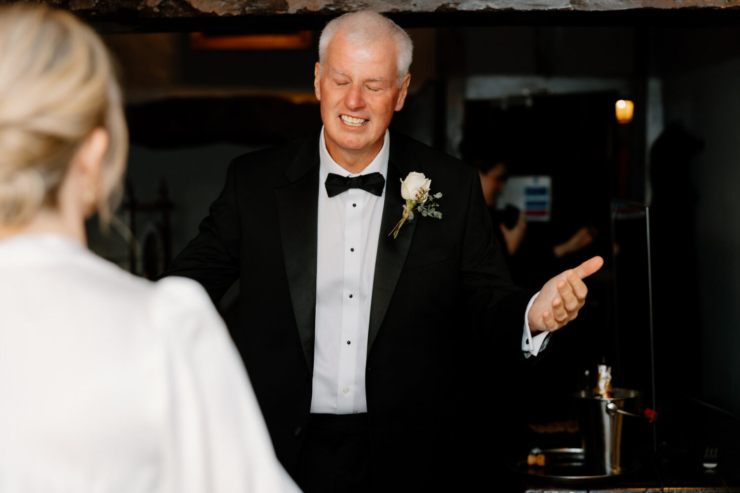 Bride's father, in a tuxedo with a boutonniere, gestures with both hands while standing indoors. A blurred person appears in the foreground.