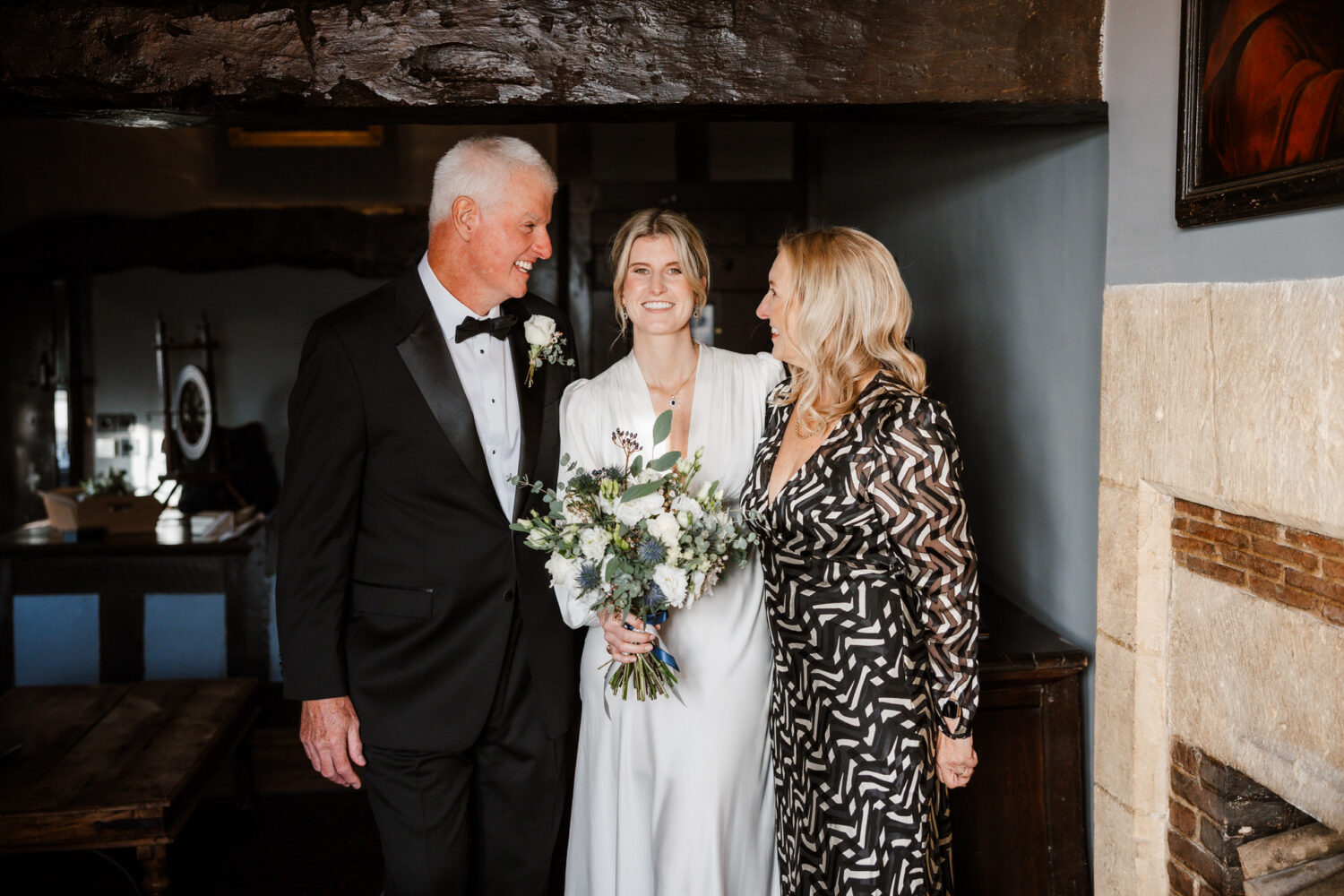 A bride stands between her father in a tuxedo and her mother in a patterned dress, holding a bouquet. They are indoors, smiling and looking towards each other.