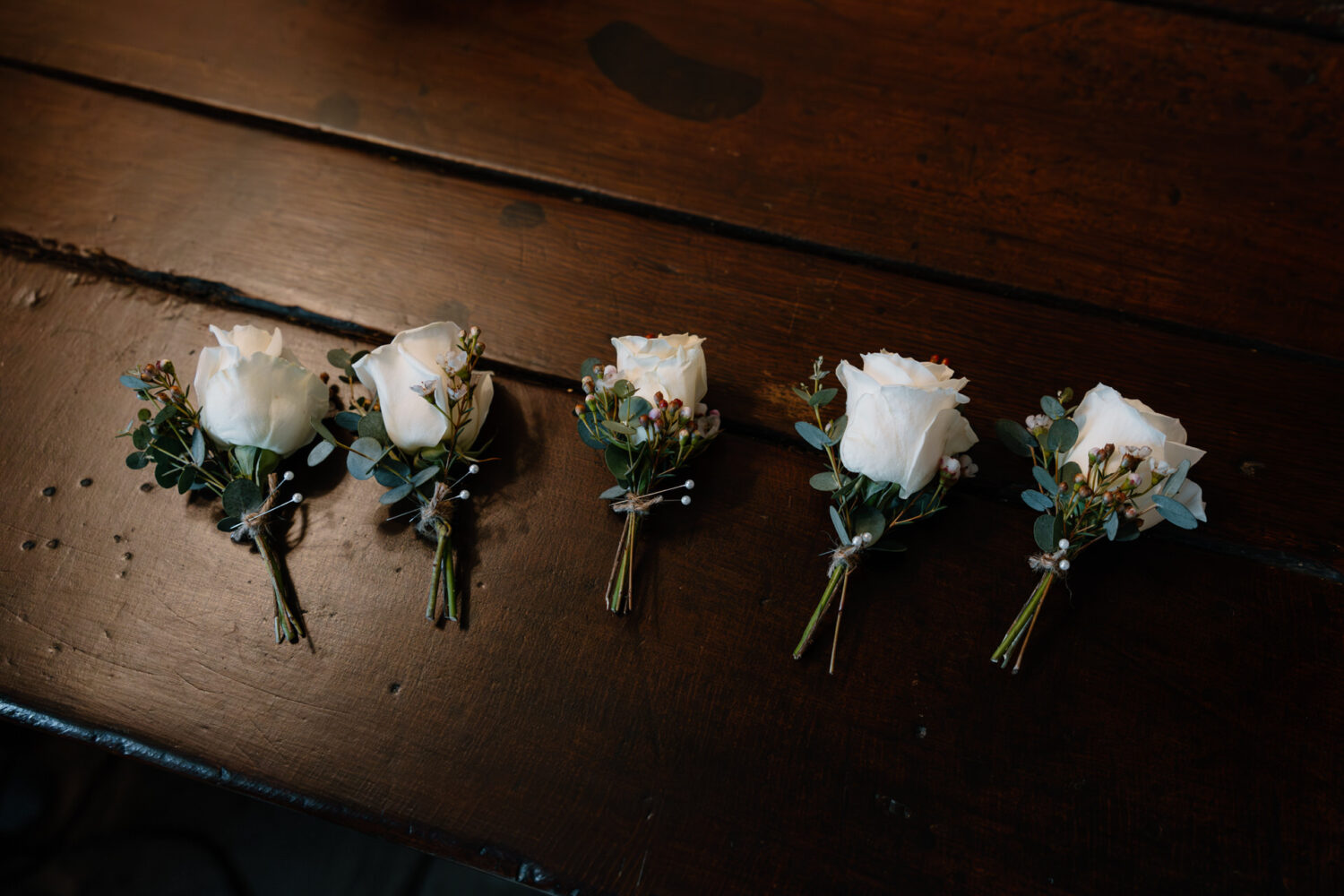 Five white rose buttonholes with greenery and small flowers are arranged on a wooden surface.