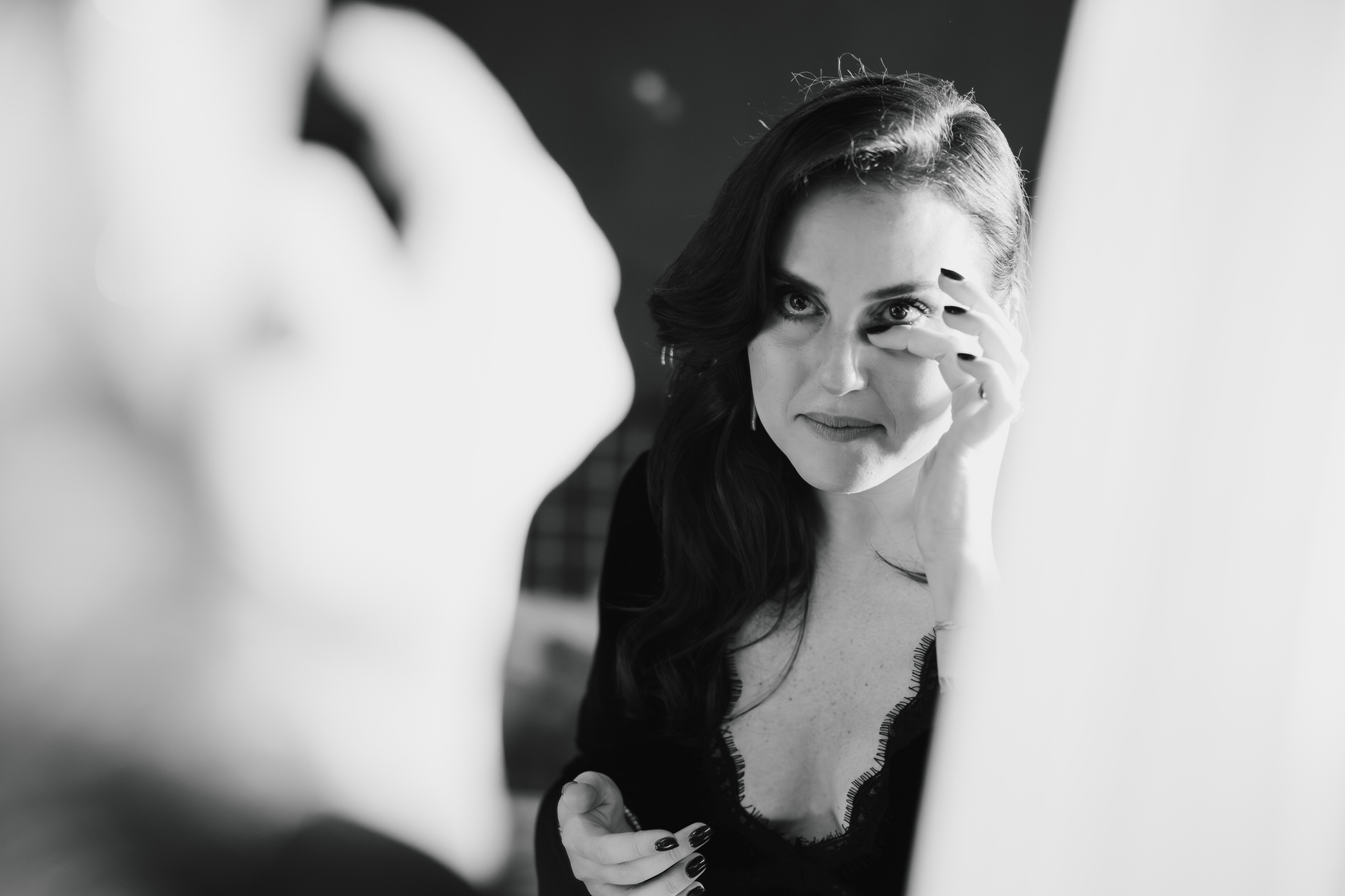 The bride's friend, in a dark top, applies makeup while looking in a black-and-white mirror.