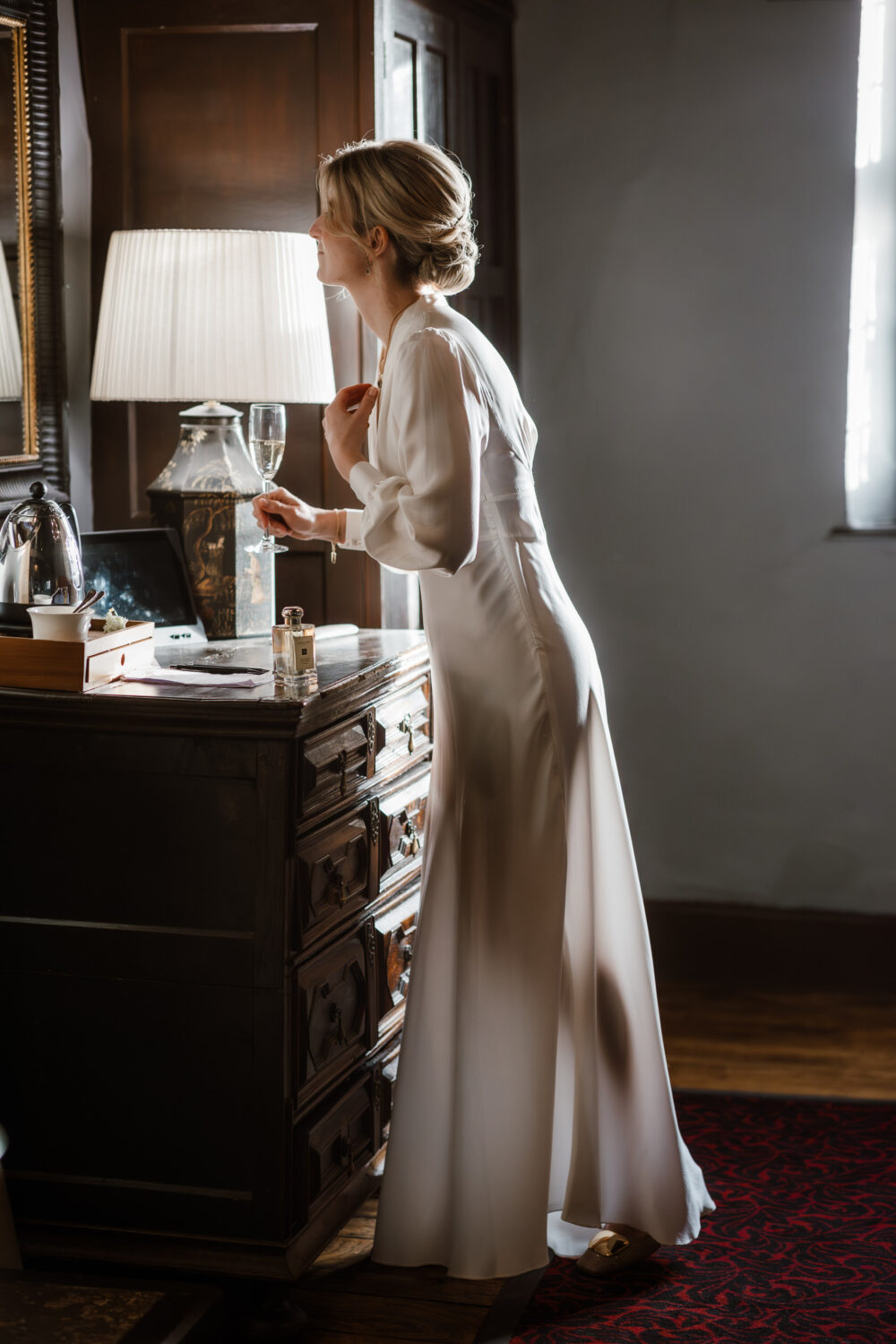 The bride in a long white dress stands by a lamp-lit dresser, looking into a mirror, holding a wine glass.