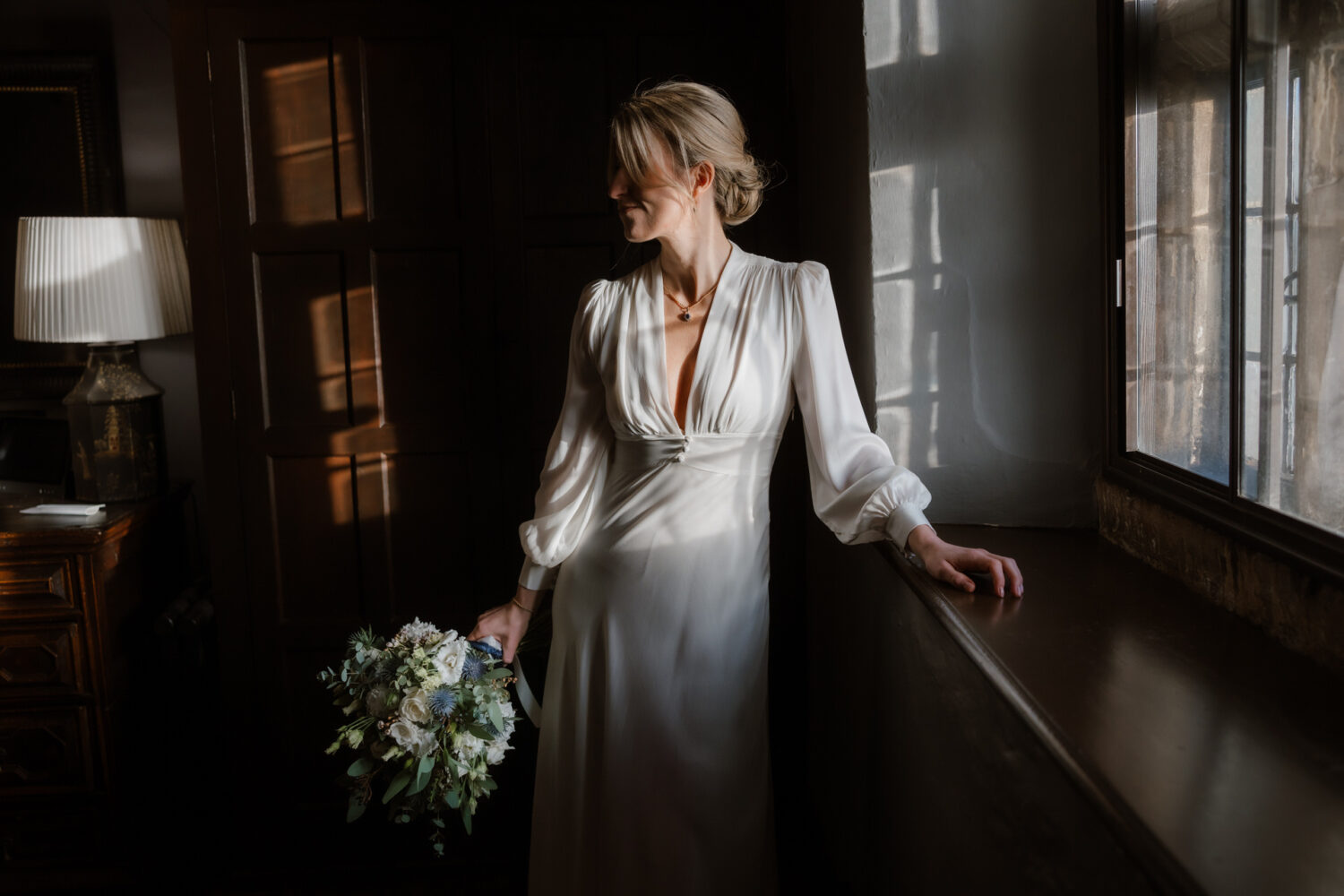 The bride is in a white dress, holding a bouquet, and stands by a dimly lit window.