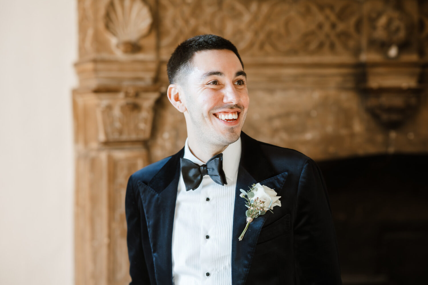 In a tuxedo with a bow tie and boutonniere, the groom smiles in front of an ornate fireplace.