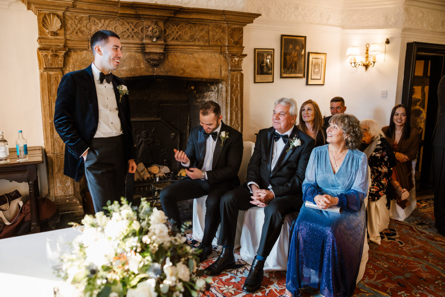 A groom in a tuxedo stands by a fireplace, speaking to seated guests in formal attire. A floral arrangement is visible in the foreground.