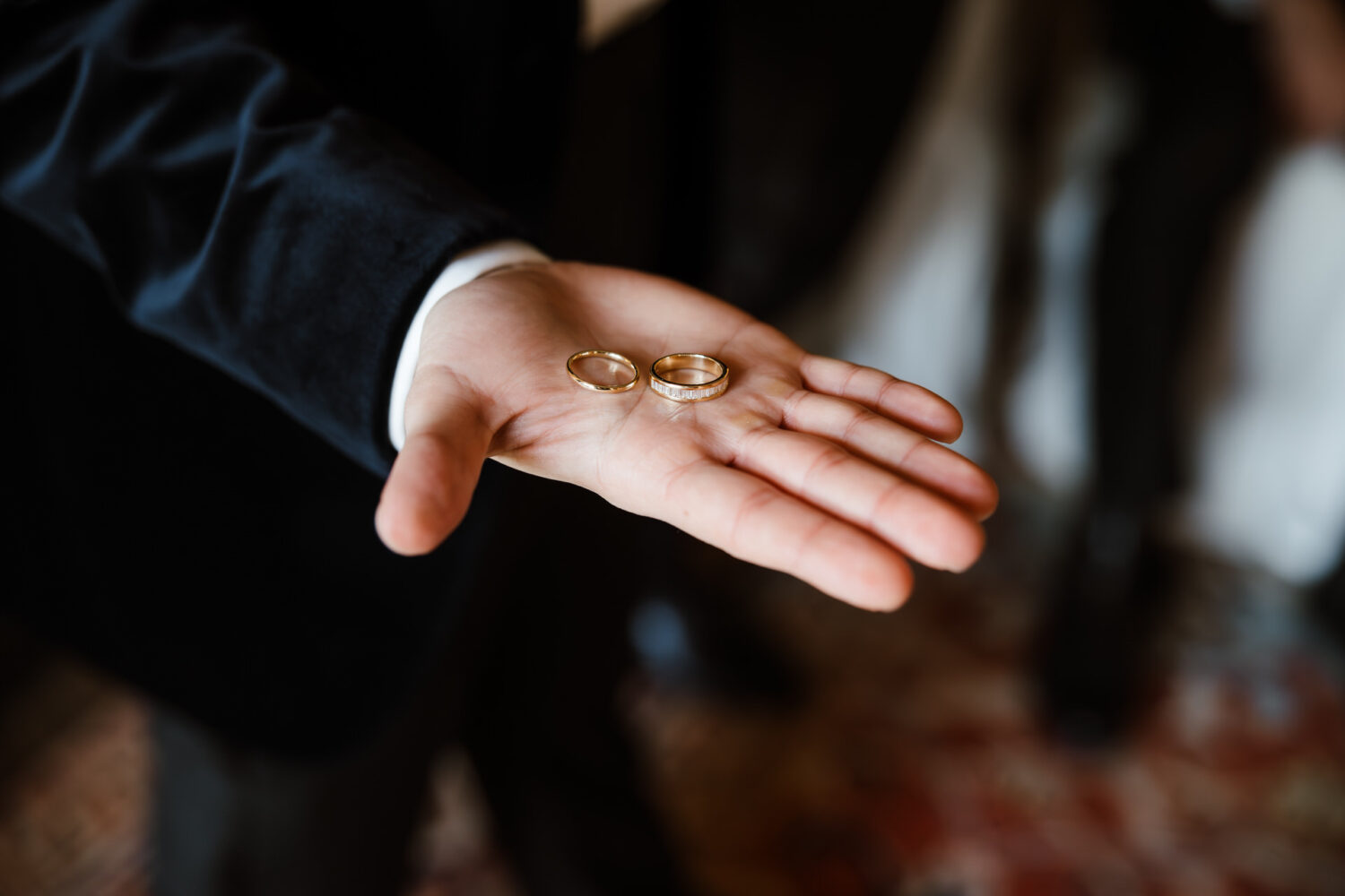 The groom in a black suit holds two gold wedding rings in their open palm.