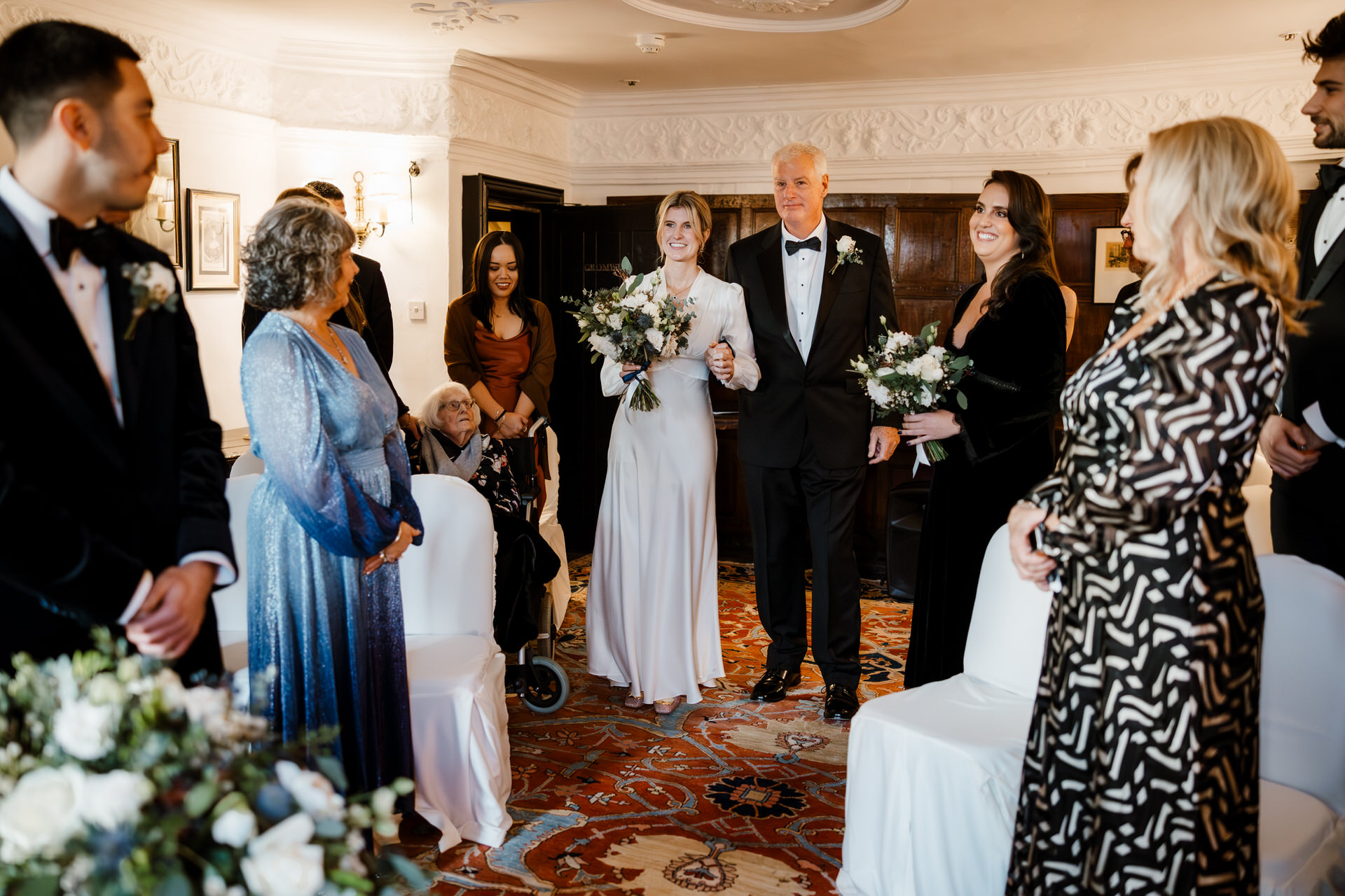 Her father escorted The bride down the aisle, surrounded by guests standing and seated in a decorated room.