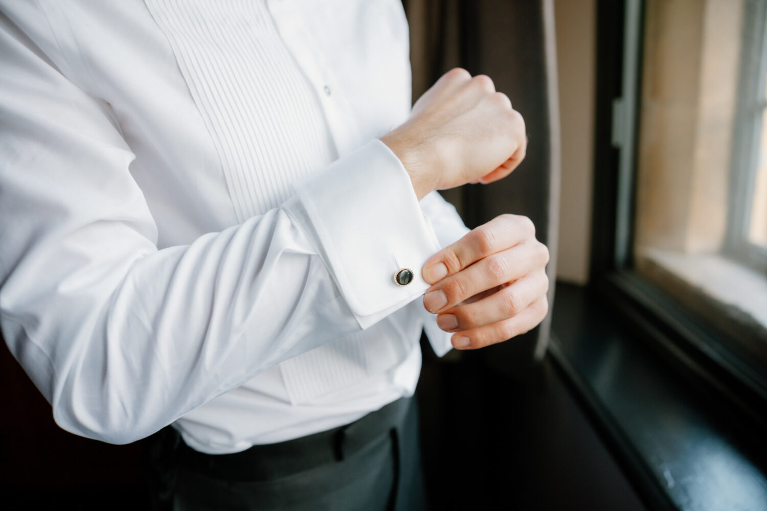 The groom in a white dress shirt adjusts cufflinks beside a window.