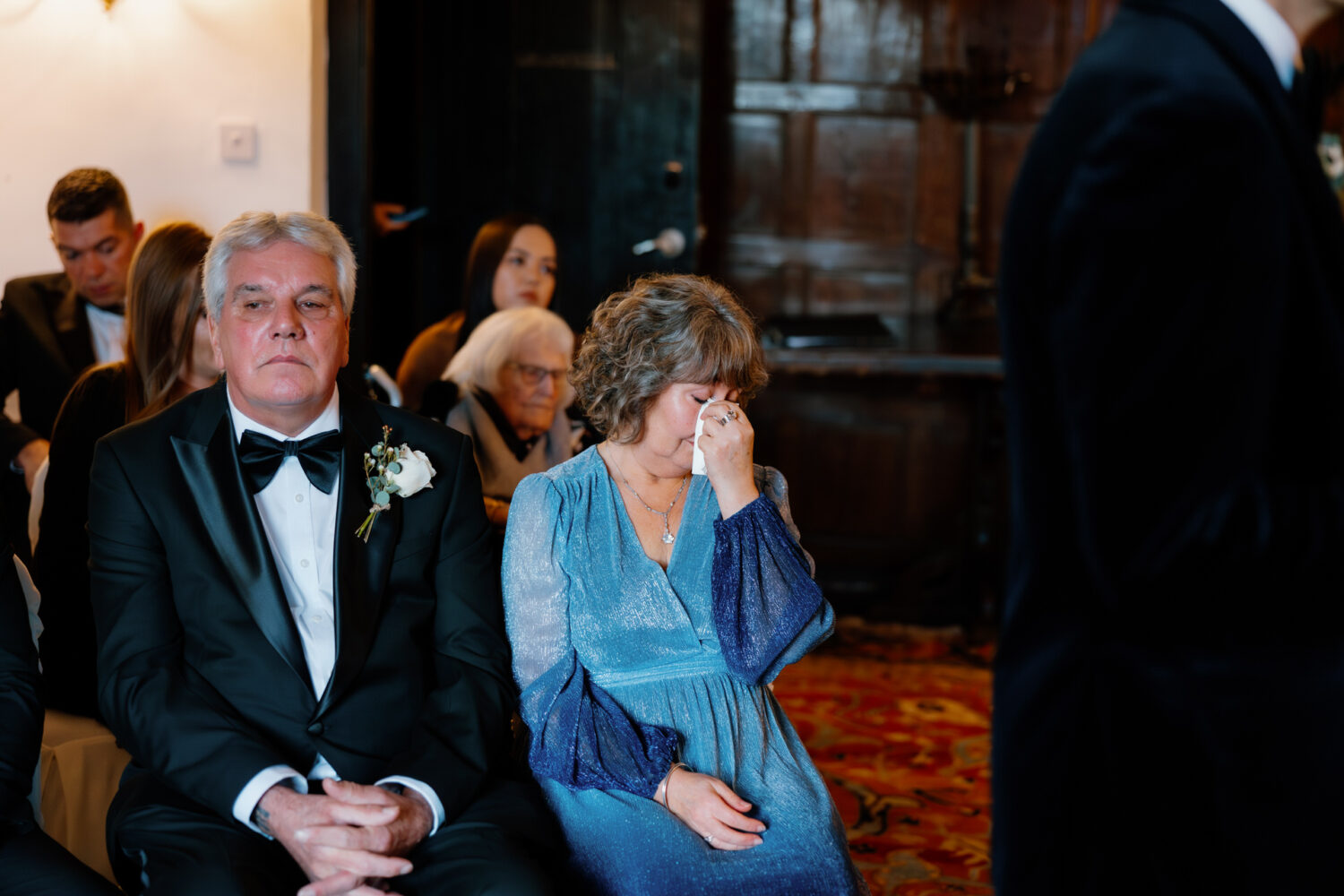 An older man in a tuxedo sits beside a woman in a blue dress who wipes her tears, surrounded by other seated attendees at a formal event.