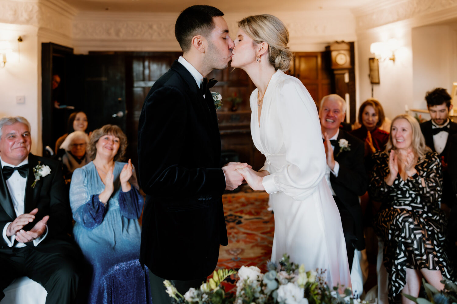 A couple kissing during their wedding ceremony, holding hands, surrounded by seated guests clapping and smiling in a warmly lit room.