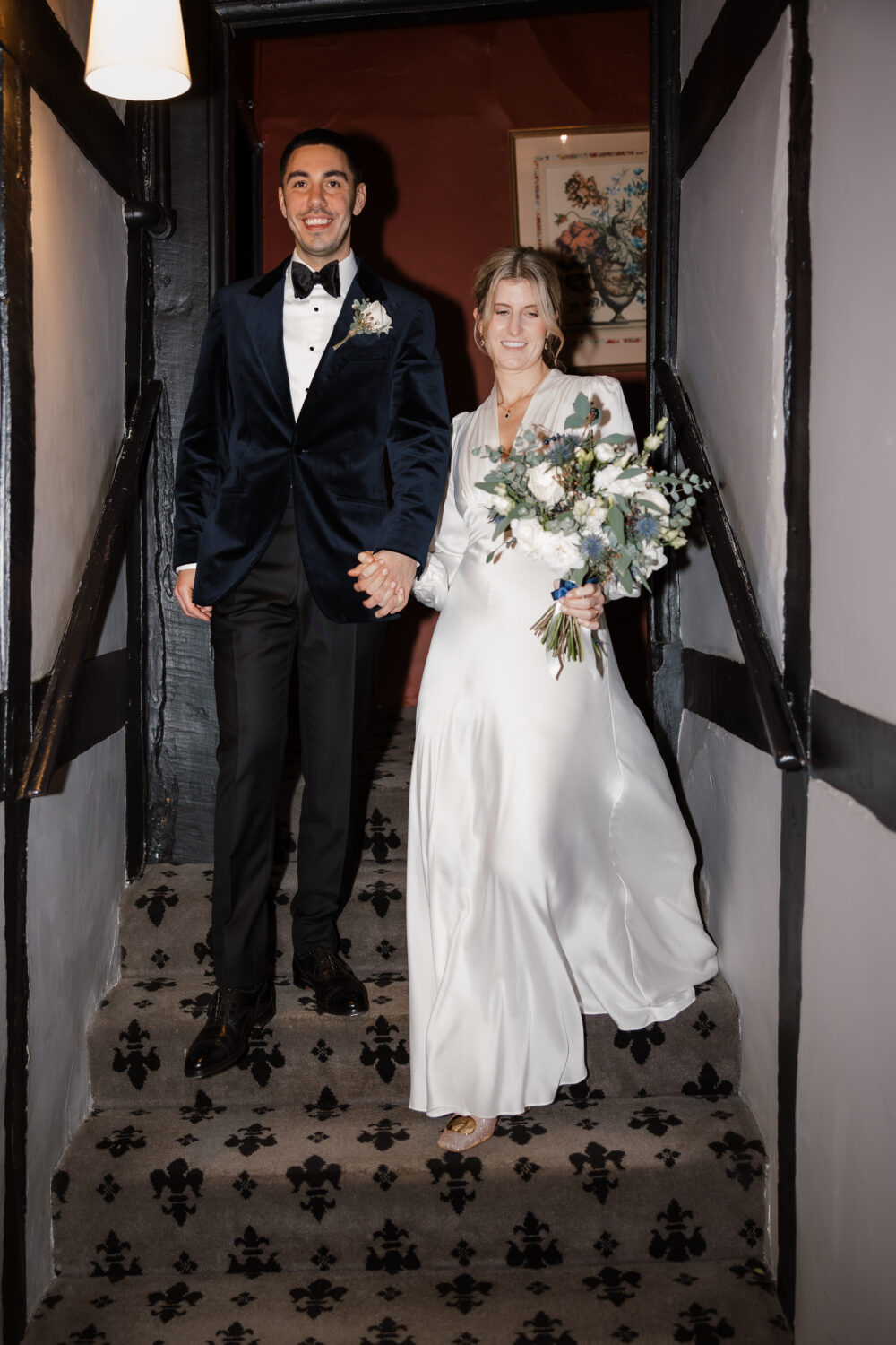 A couple in formal attire descends a staircase; the man wears a dark suit, and the woman in a white gown holds a bouquet.