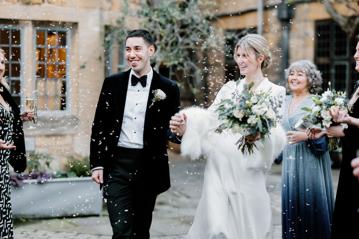 A smiling couple exits a building, surrounded by people and falling confetti. The woman holds a bouquet, and the man wears a black suit.