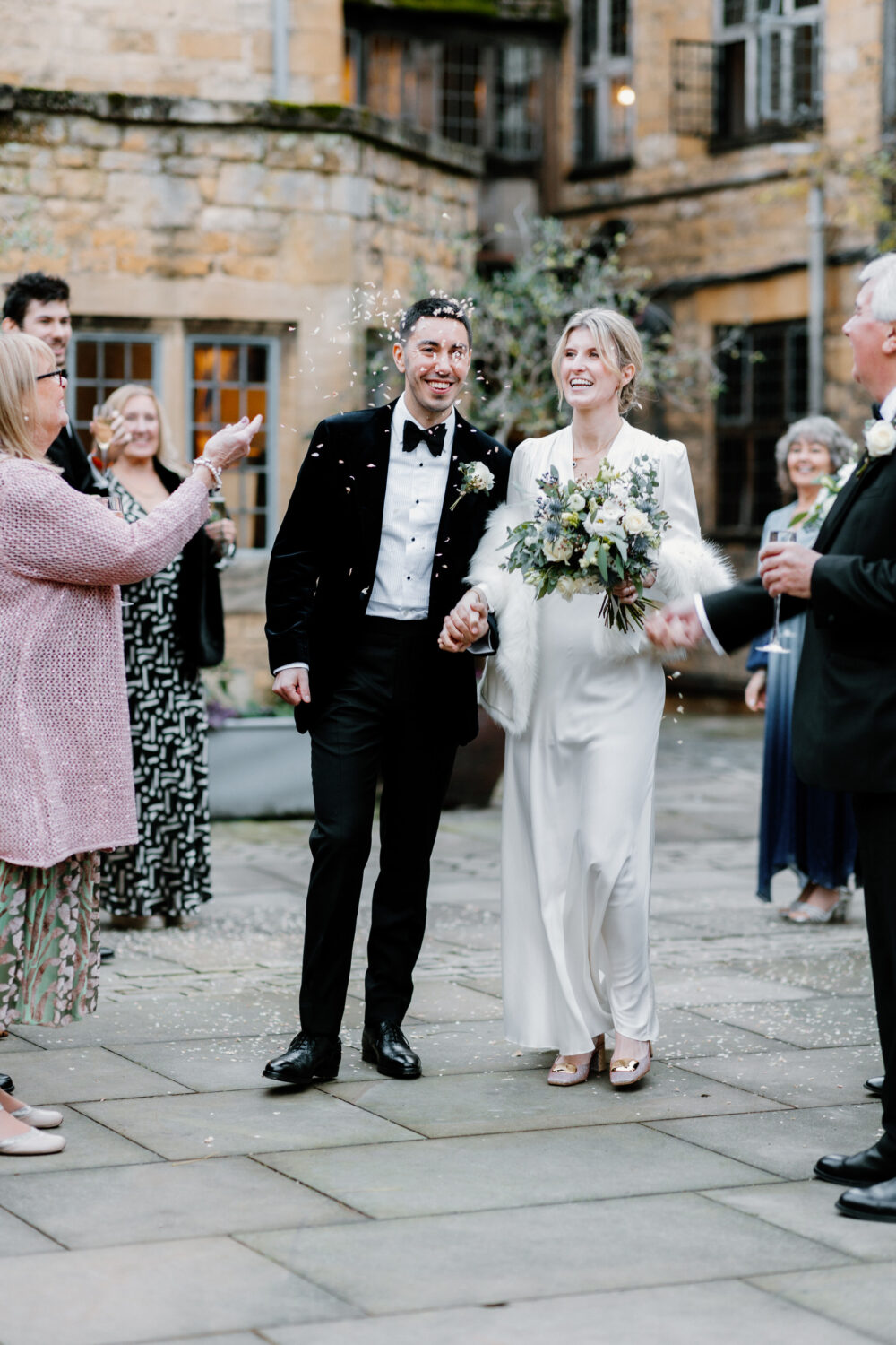 A bride and groom walk hand in hand outdoors, smiling as guests toss confetti. The bride wears a white dress and holds a bouquet; the groom is in a tuxedo. Stone buildings are in the background.