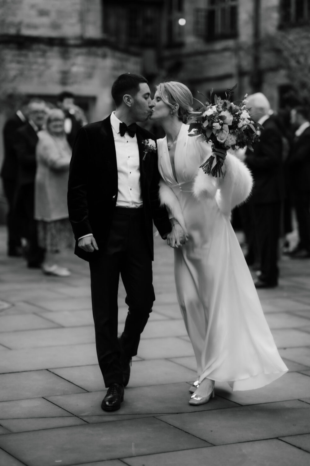 Bride and groom share a kiss while walking outside. The bride holds a bouquet and wears a white dress with a fur stole, while the groom wears a dark suit with a bow tie.