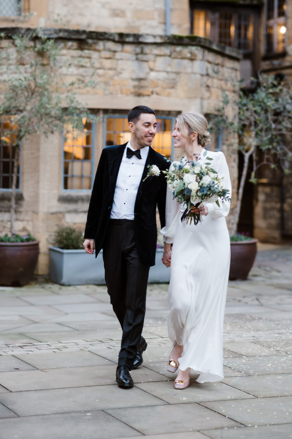 A couple in formal attire walks hand in hand outdoors. The woman holds a bouquet and wears a white dress, while the man wears a black tuxedo. They appear to be smiling at each other.