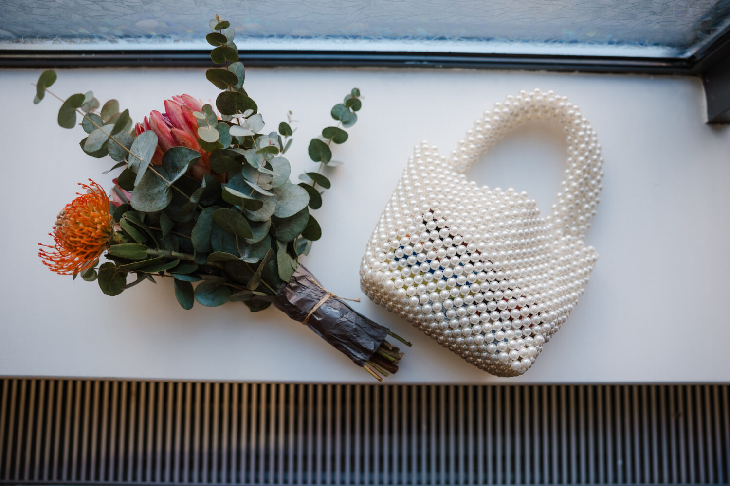 A bouquet of colorful flowers lies beside a small, pearl-beaded handbag on a white surface.