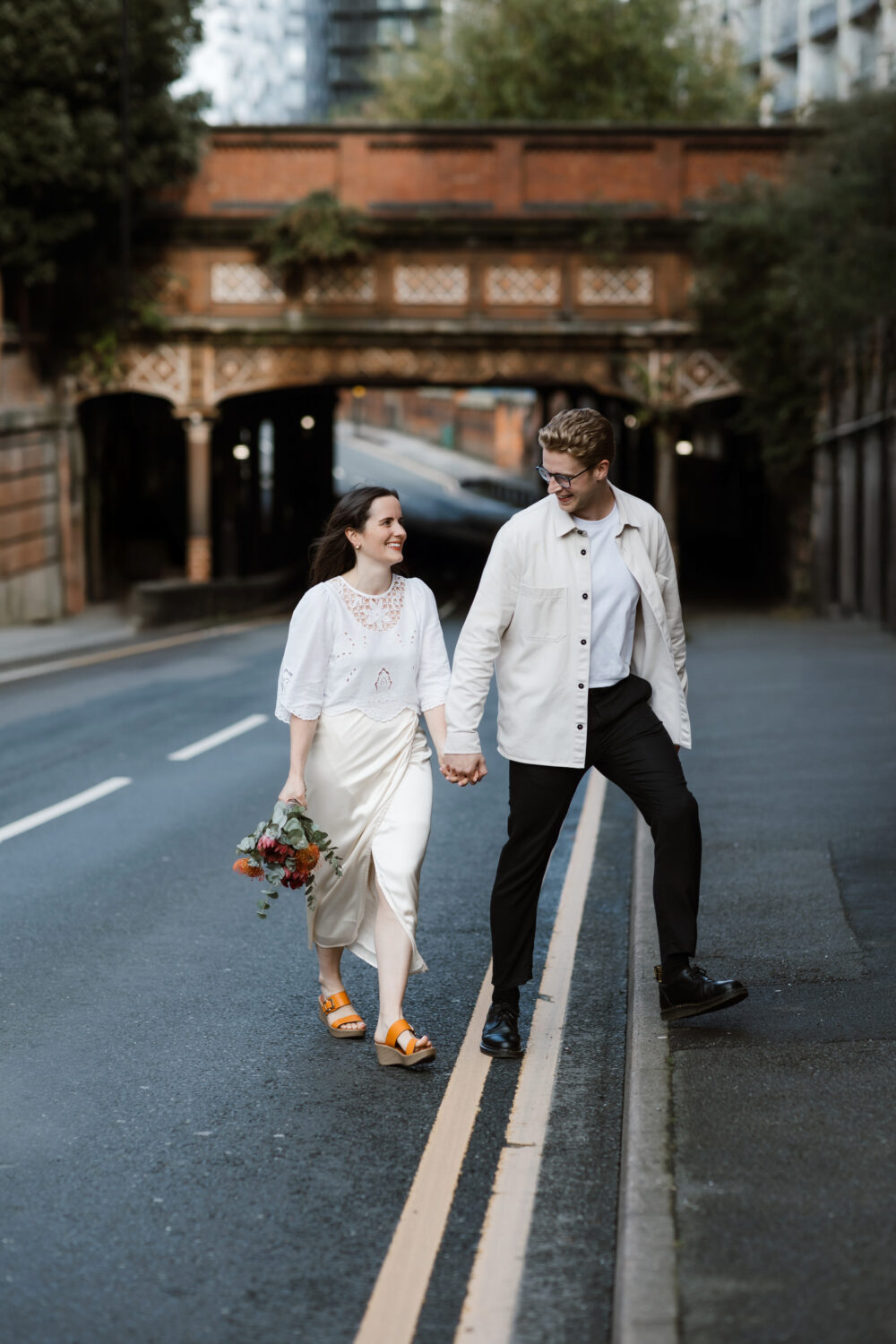 The bride and groom stroll hand in hand down a Birmingham street, with the groom stepping onto a curb. The bride gracefully holds a bouquet. A decorative brick bridge serves as the backdrop for their charming civil wedding moment.