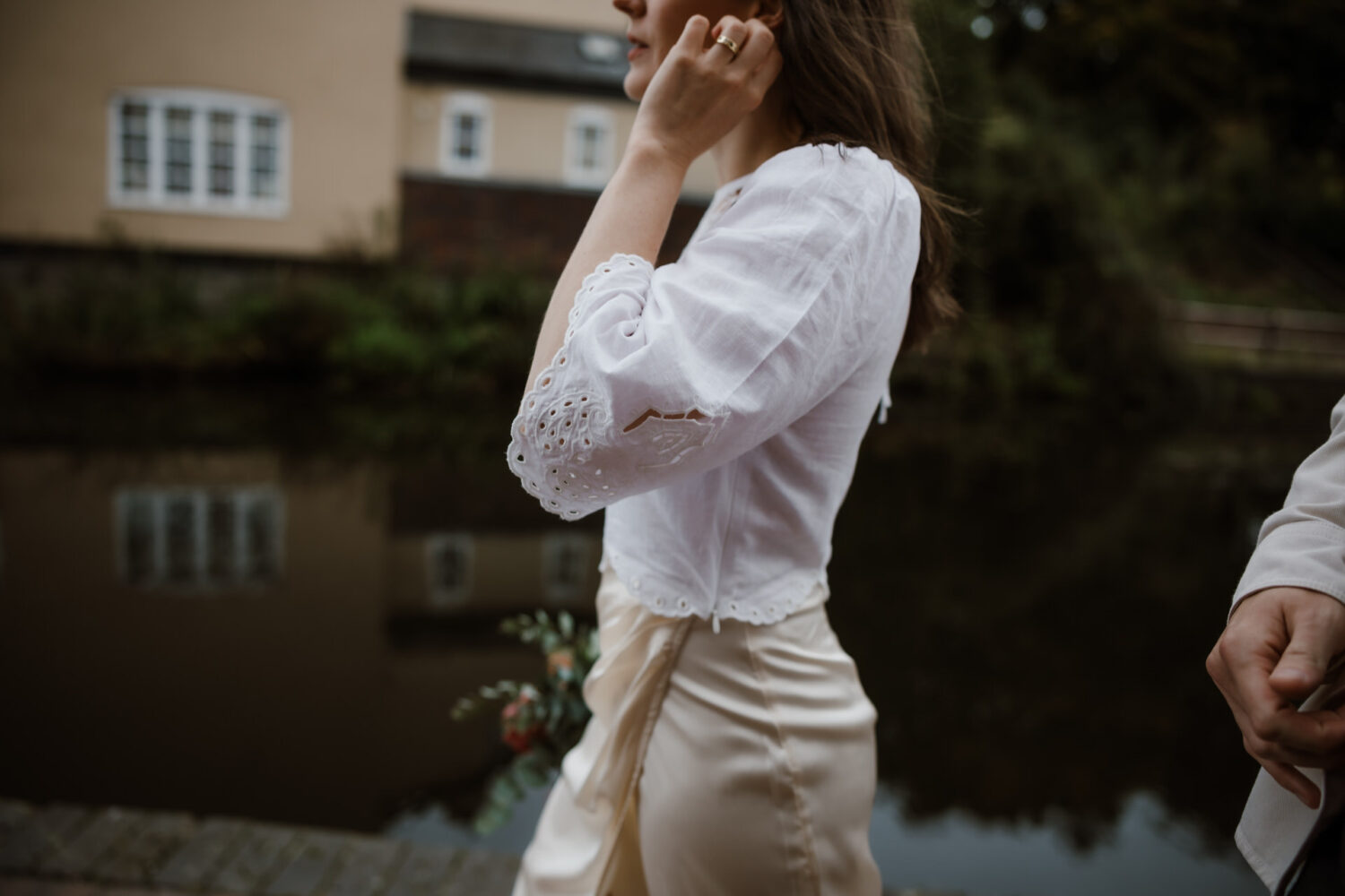 Person in a white blouse and beige skirt holding a small bouquet while walking by a canal.