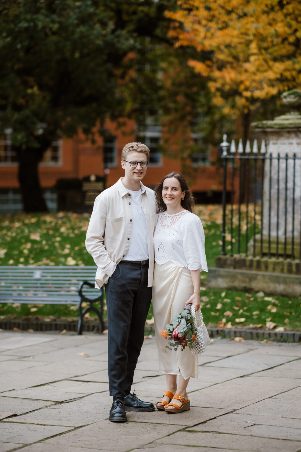 A bride and groom stand together outdoors, dressed in casual yet formal attire, embodying the spirit of a Birmingham civil wedding. The bride holds flowers, adding a touch of elegance against the backdrop of autumn trees and a nearby bench.