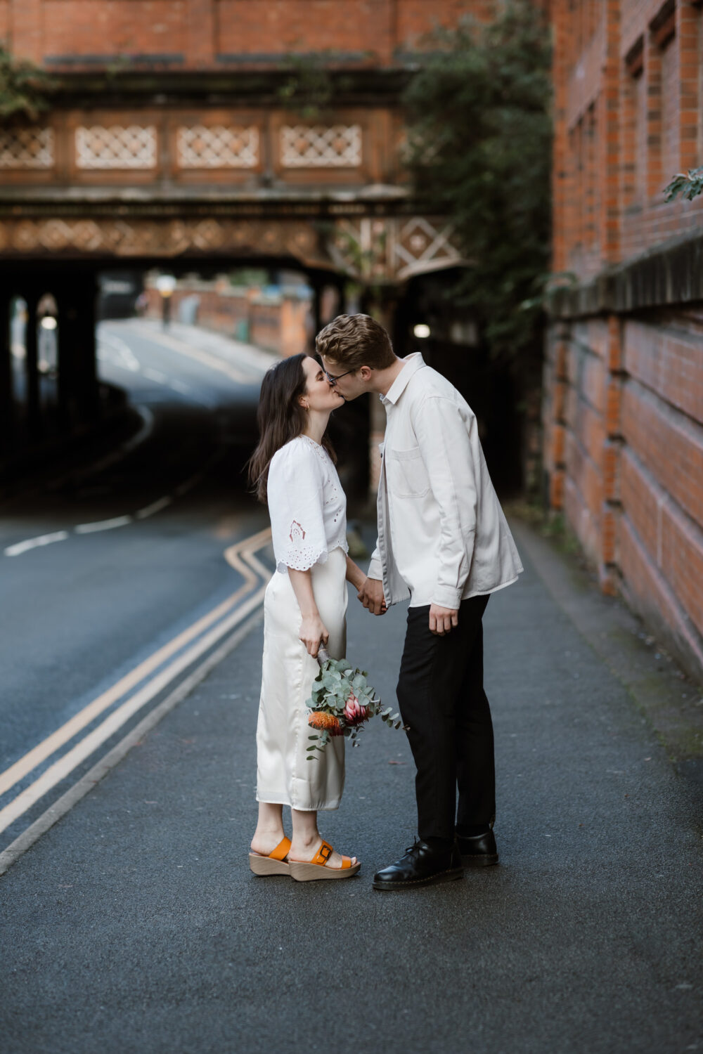 In Birmingham, the bride and groom share a tender kiss on the sidewalk next to a bustling road, their hands entwined and holding a bouquet of flowers. A brick wall and bridge stand gracefully in the background, framing this intimate moment from their civil wedding day.