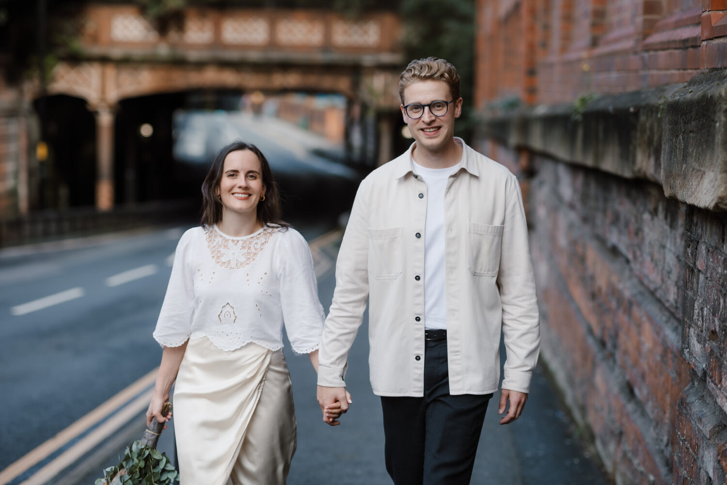 The bride and groom, hand in hand, stroll along a Birmingham street, with a brick wall and bridge framing the background. She glows in her white dress, while he sports a light jacket. Both are smiling radiantly.