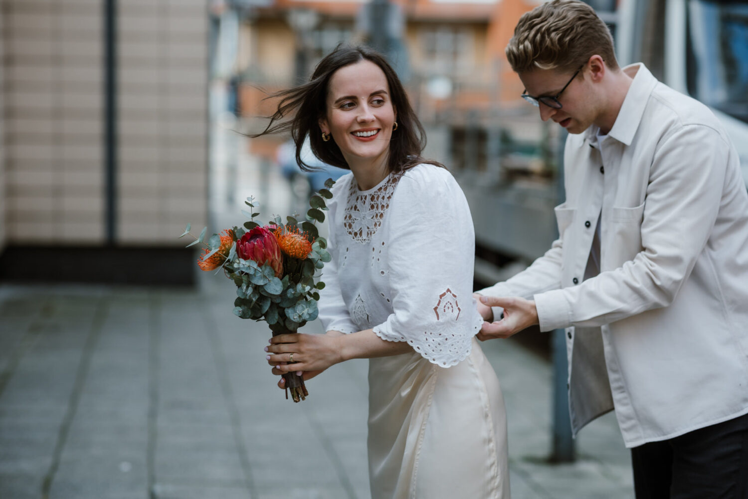 A woman holding a bouquet smiles while walking as her newlywed husband adjusts her sleeve on a city street. Both wear light-colored clothing.