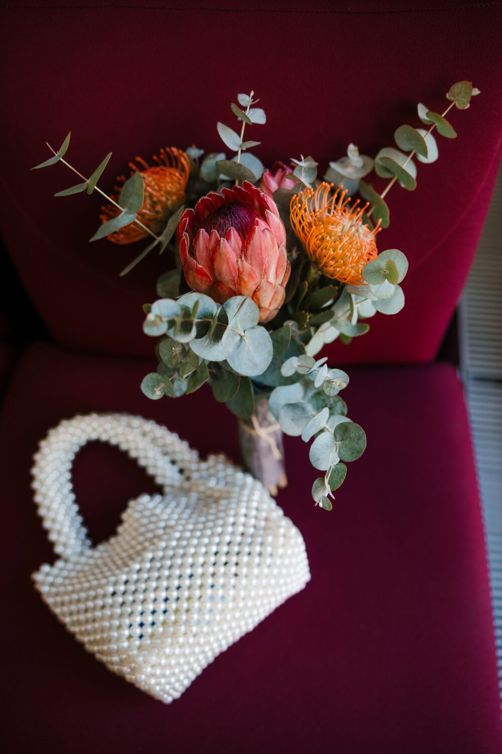 A bouquet with red and orange protea flowers and green eucalyptus rests on a maroon chair. A white beaded handbag is placed beside it.