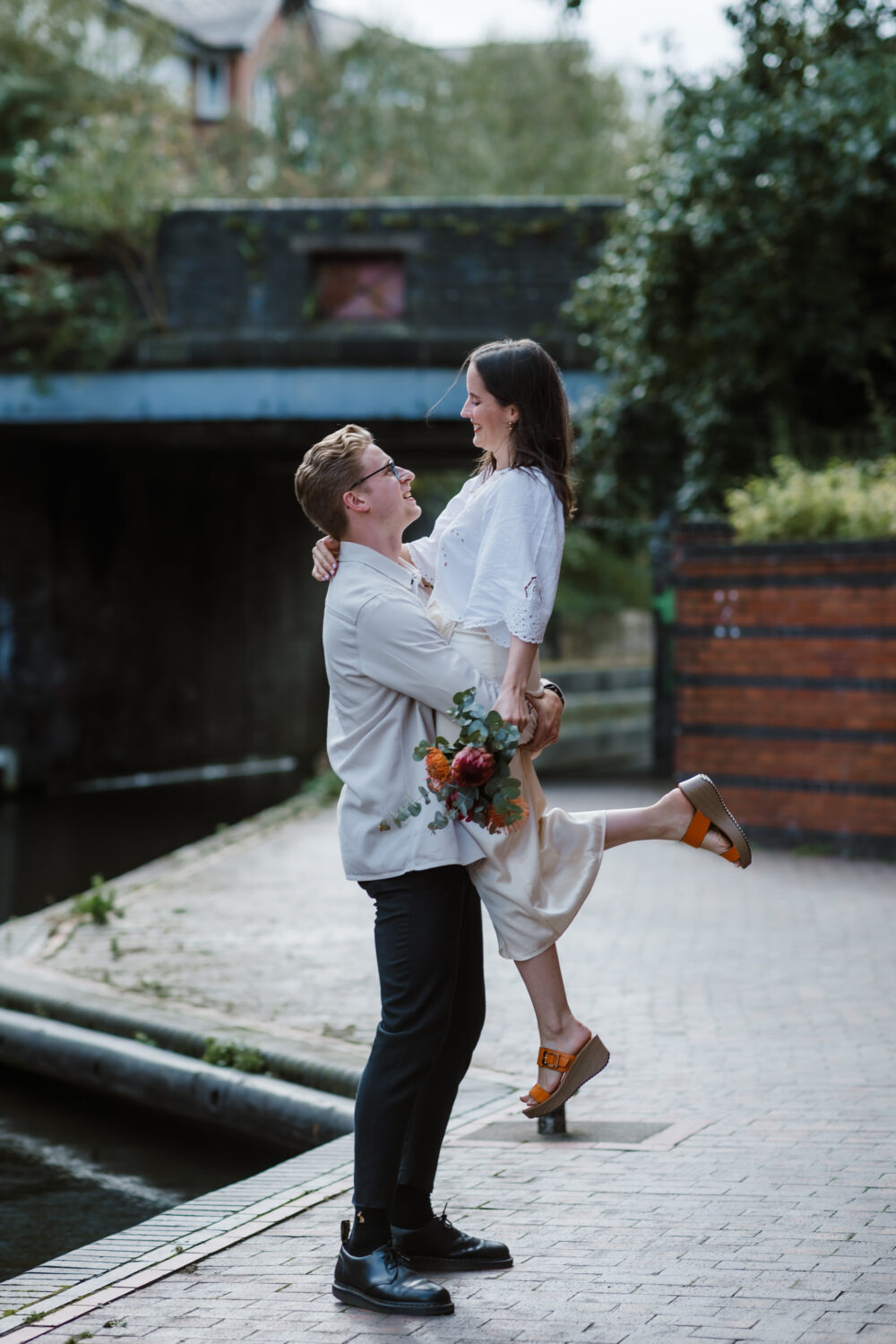 A person lifts another person joyfully near a canal. The standing person holds a bouquet of flowers.