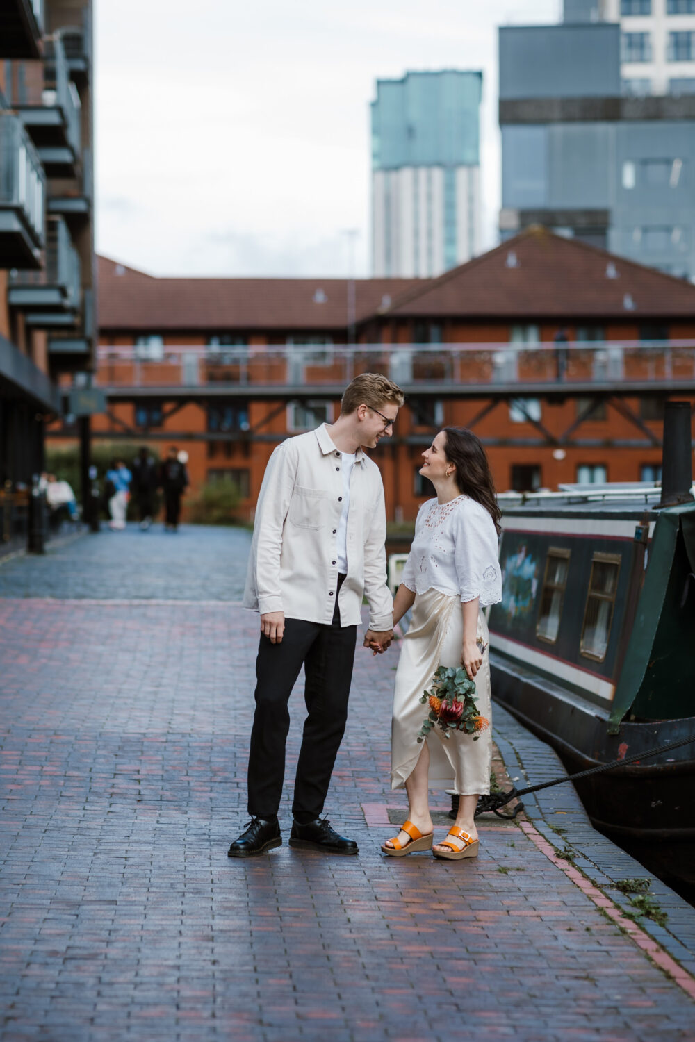 A couple stands holding hands on a brick pathway by the Birmingham canals, reminiscent of a bride and groom's special moment. They are facing each other and smiling, with urban buildings rising in the background.