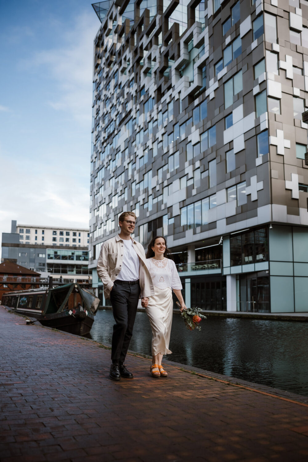 The bride and groom walk along a canal path beside The Cube Birmingham, its modern architecture providing a striking backdrop. The groom wears a light jacket and trousers, while the bride, in her white dress and sandals, holds a small bouquet of flowers after their civil wedding.