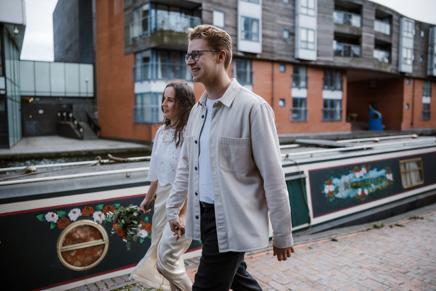 A couple, resembling the bride and groom, strolls along a canal path beside a narrowboat, against the backdrop of modern buildings. The man wears glasses and a light-colored jacket, while the woman holds a bouquet.