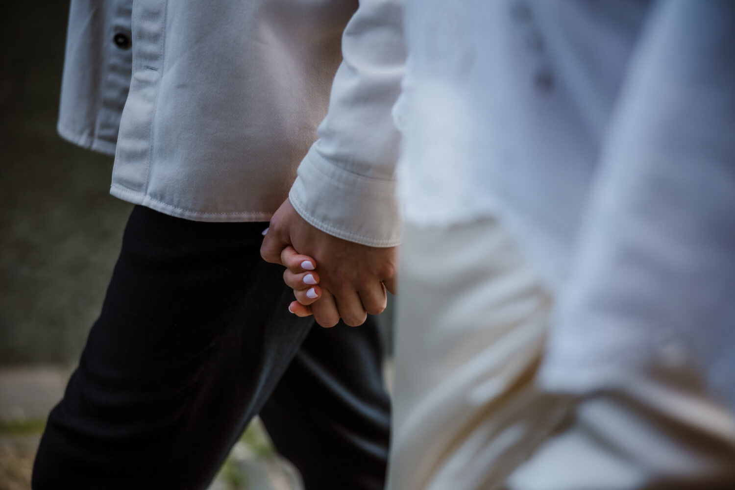 Two people are holding hands, with one wearing dark pants and the other in light clothing. The focus is on their hands.