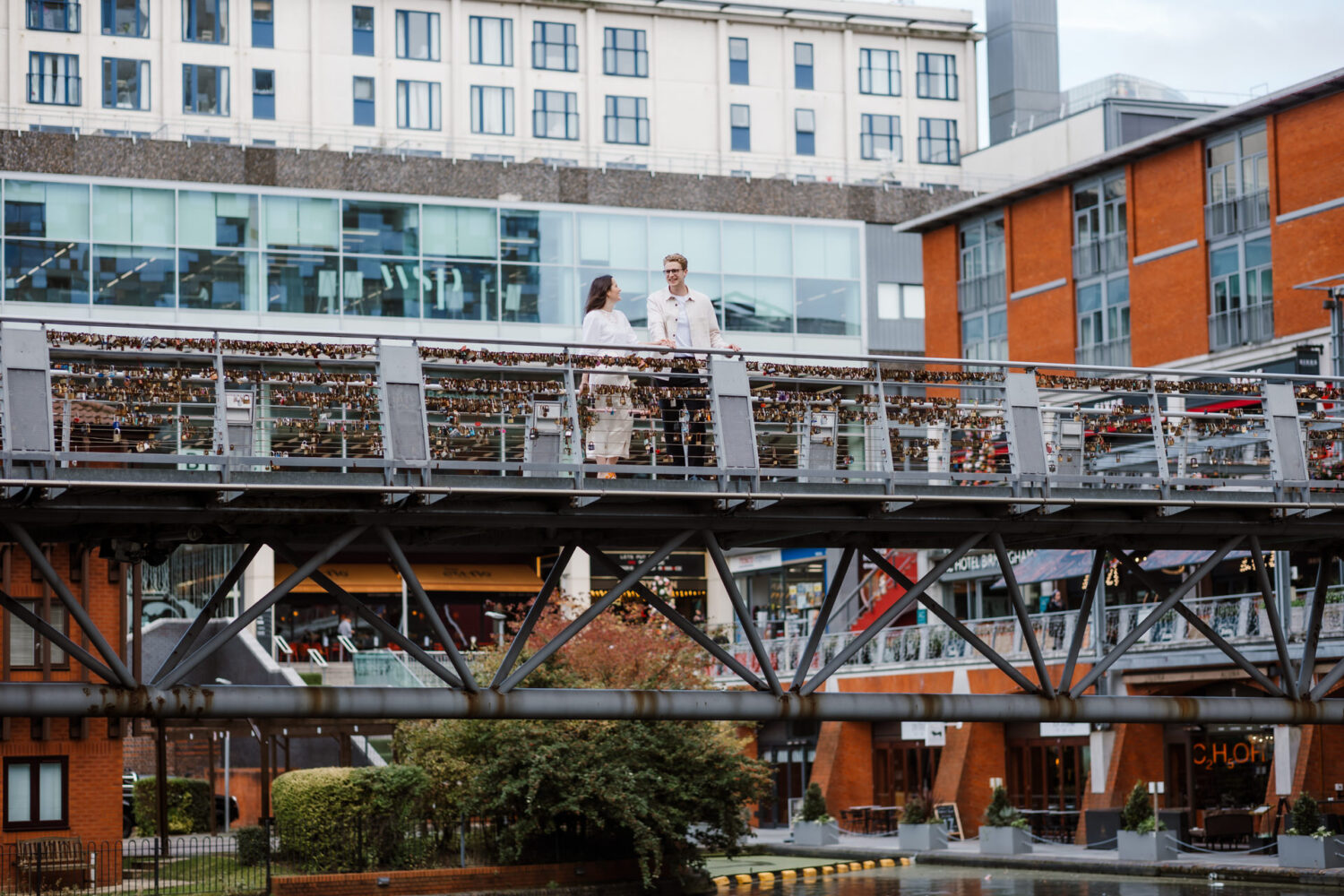 The bride and groom stand on a pedestrian bridge adorned with love locks, surrounded by the modern buildings and greenery of Mailbox Birmingham, celebrating their civil wedding.