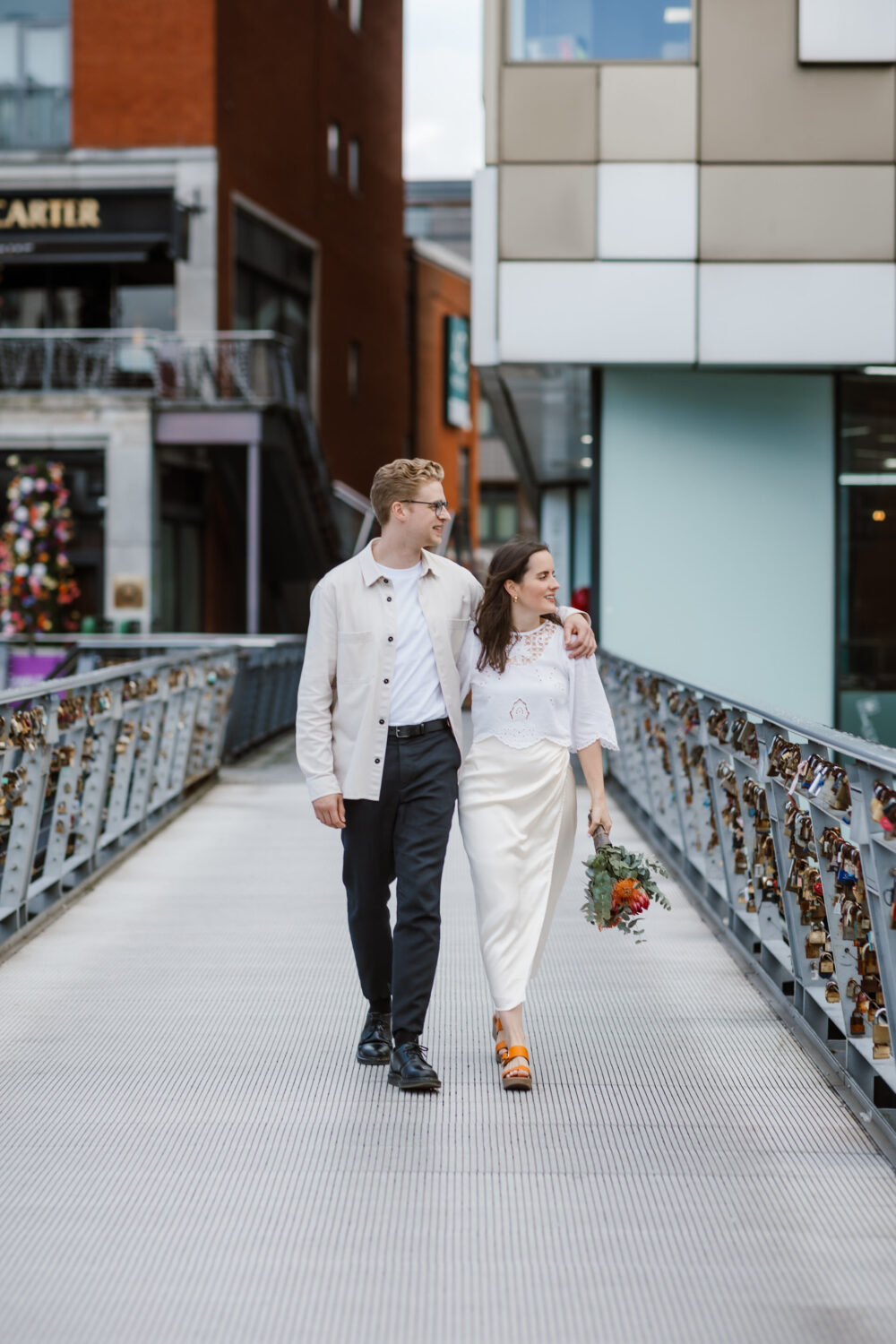 A couple walks arm-in-arm on a bridge lined with love locks. The man wears a light jacket, and the woman holds a bouquet of flowers, wearing a white outfit. Urban setting is visible in the background.