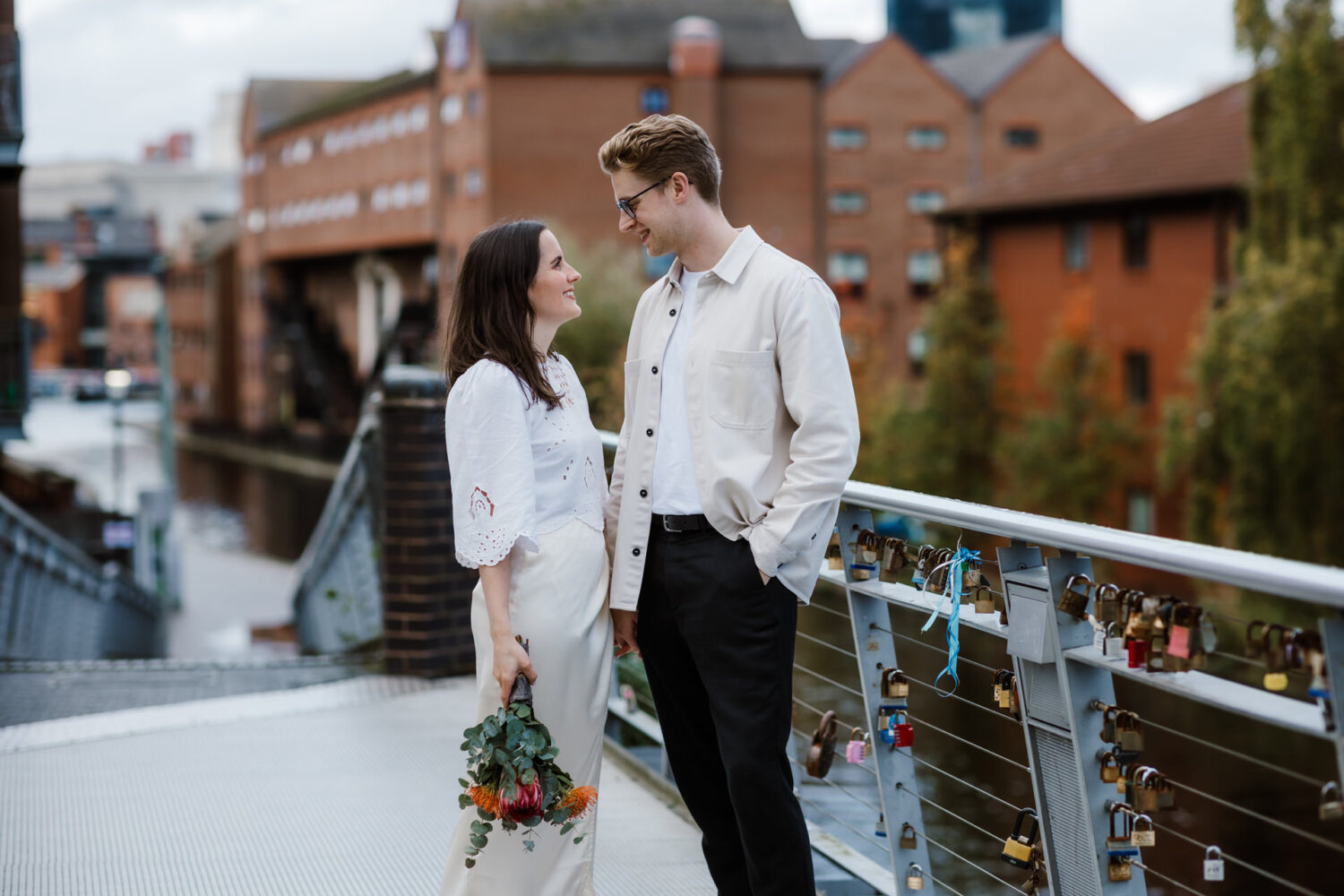 The bride and groom stand on a bridge adorned with love locks, facing each other and smiling. The woman holds a bouquet, capturing the essence of their Birmingham civil wedding. Red brick buildings provide a charming backdrop to this romantic moment.