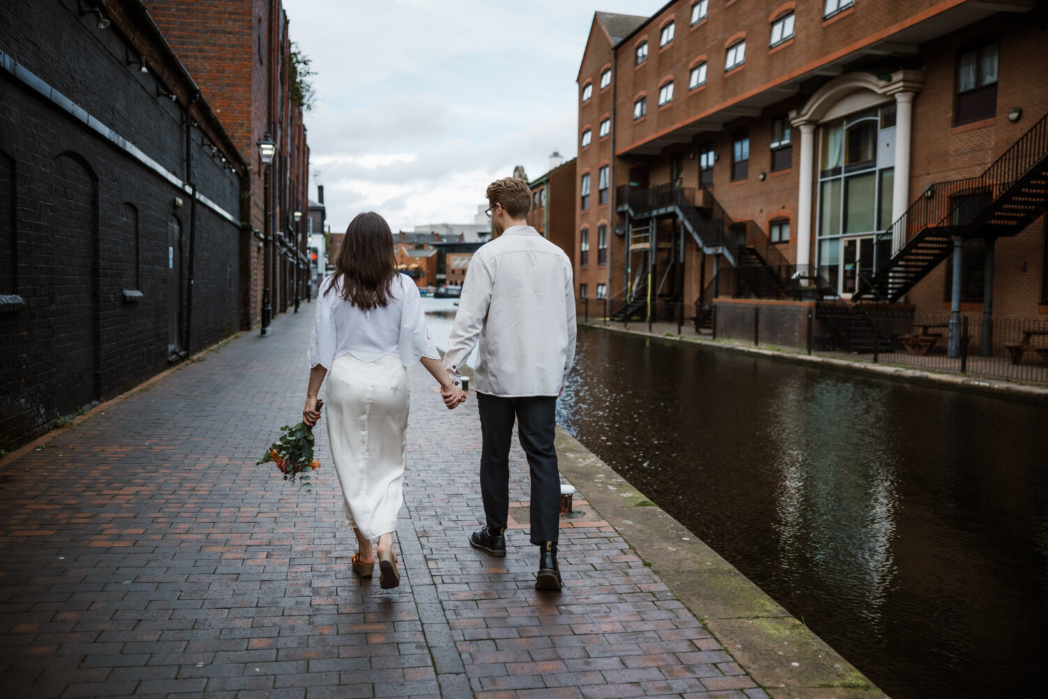 A couple walking hand in hand along a canal-side brick path, with the woman holding a bouquet of flowers.