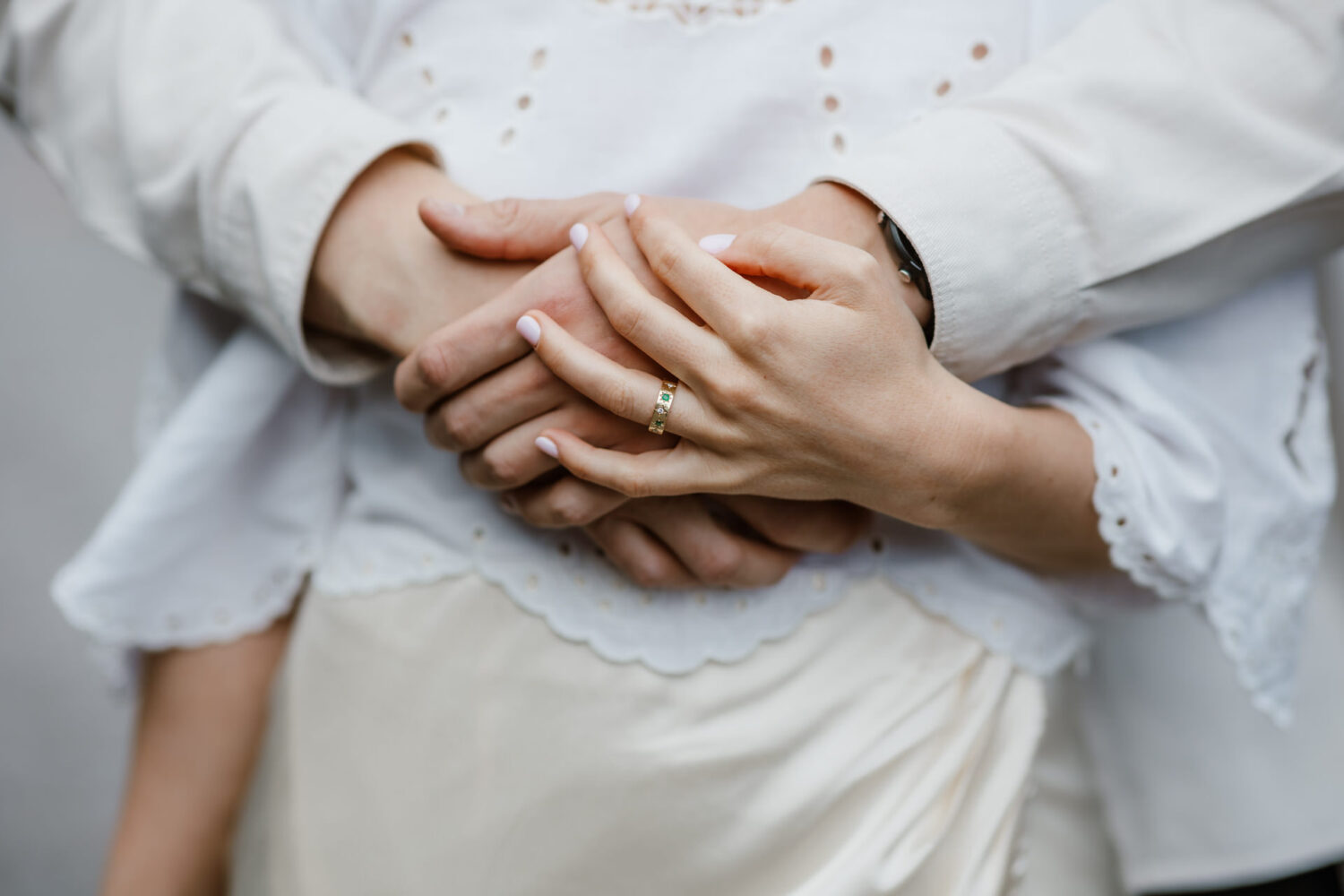 Two people stand close, hands interlocked. The front person wears a ring on the left hand. Both wear light-colored clothing.