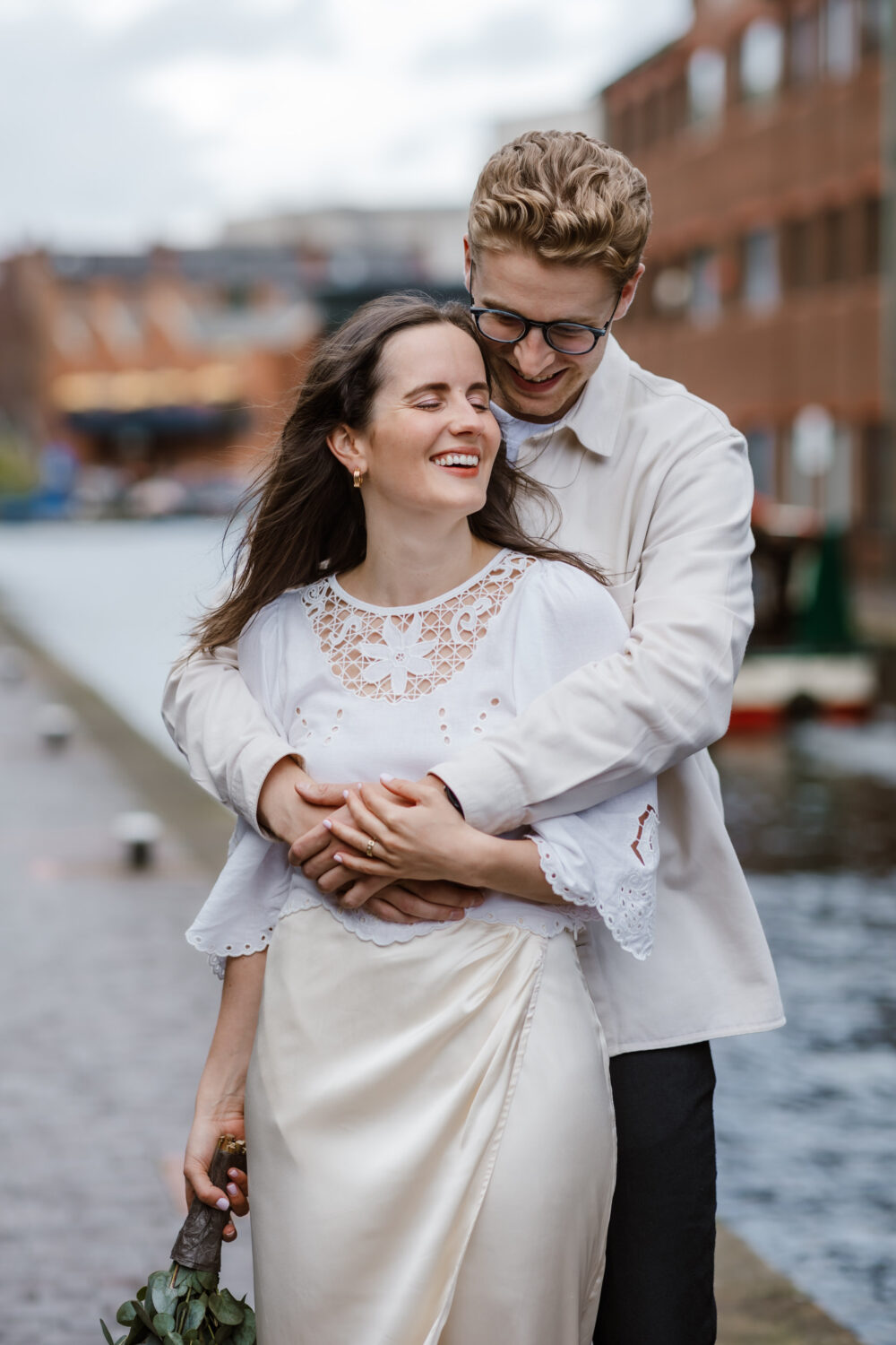 A couple smiling and hugging by a canal. The woman holds a bouquet of flowers. They stand on a cobblestone path with brick buildings in the background.