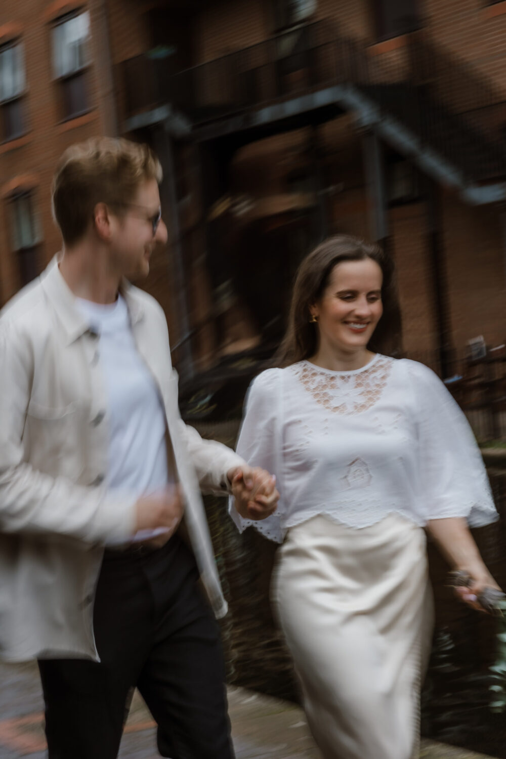 A man and woman walk hand in hand, smiling. They are outside near a brick building. The image is slightly blurred, suggesting motion.