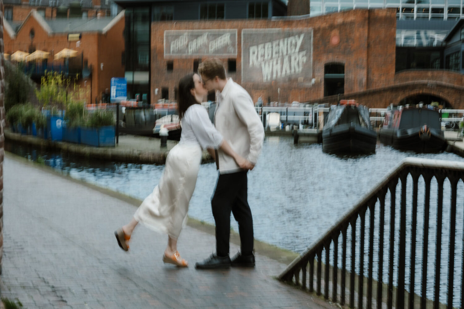 A couple kissing by a canal, with brick buildings and narrowboats in the background. The image is slightly blurred.