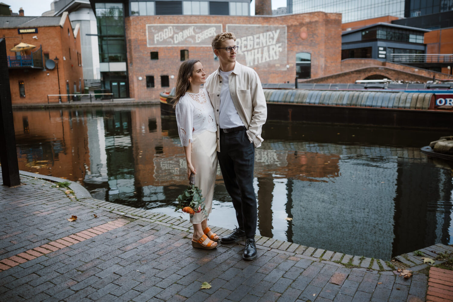 A couple stands by a canal, with brick buildings in the background. The woman holds orange flowers, and the man looks away. The scene reflects an urban waterfront area.