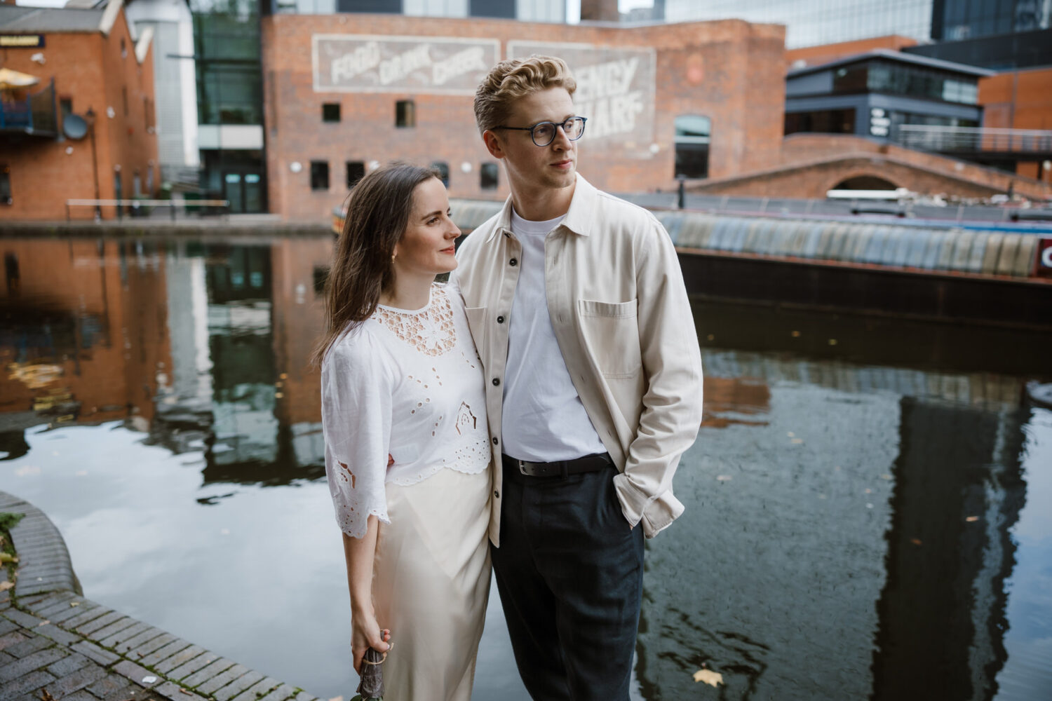 The bride and groom stand by a canal, with brick buildings and a canal boat in the background. The woman is wearing a white blouse and skirt; the man is in a light jacket and glasses.