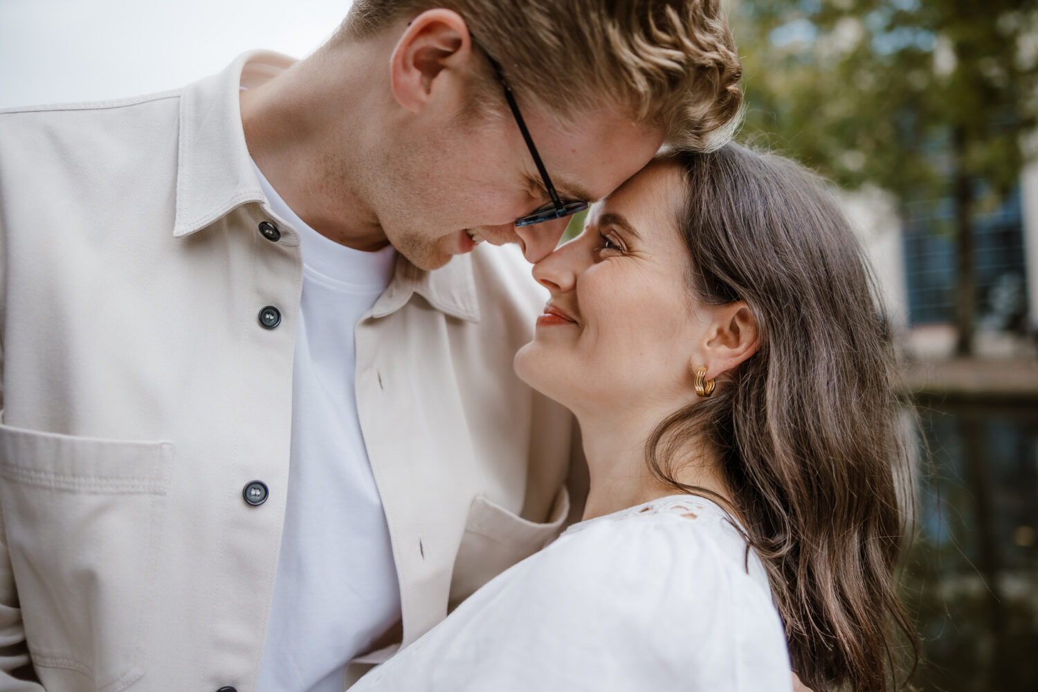 A couple stands closely, touching foreheads and smiling at each other. They are outdoors with trees and a building in the background.