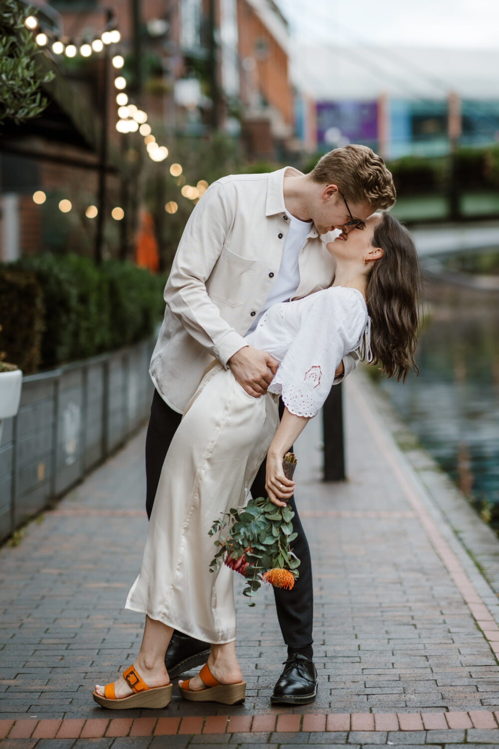 A couple kisses while the man dips the woman backwards on a brick pathway by a canal. She holds a bouquet of flowers. String lights and buildings are visible in the background.