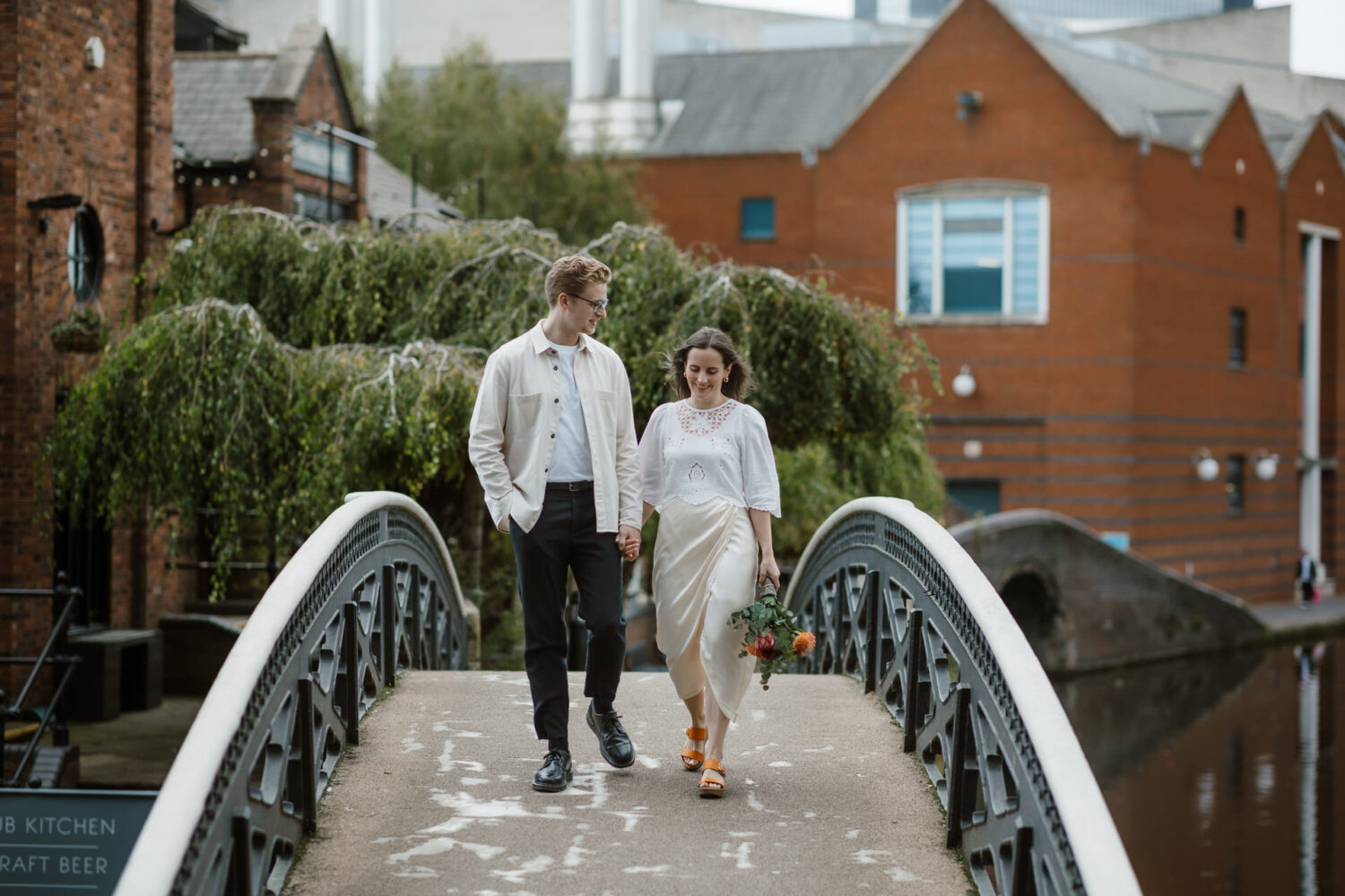 A couple holding hands and walking on a small bridge in an urban area, with buildings and trees in the background. The woman carries a bouquet of flowers.